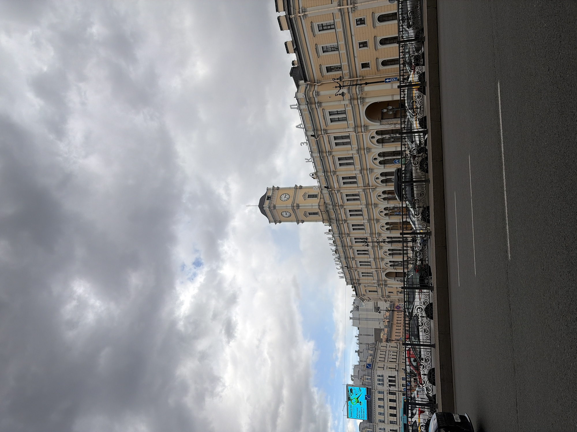 This image captures a wide street scene in Saint Petersburg, Russia, centered on the historic Moskovsky Vokzal (Moscow Railway Station) under a cloudy sky. The prominent subject is the large, multi-story building of the railway station, rendered in a light yellow or beige color, featuring classical architectural design with numerous arched windows, decorative cornices, and pilasters. A distinctive clock tower, equipped with dark-faced clocks, rises from the building's facade.
In the foreground, a wide, dark asphalt road with clear white lane markings runs parallel to the building. A decorative black wrought-iron fence separates the road from the sidewalk and the station's premises, behind which several parked vehicles, including cars and what appears to be a bus, are partially visible. The sky is largely overcast with heavy, grey clouds, though patches of brighter sky and a hint of blue are discernible, suggesting variable cloud cover during daytime.
Notable details include the architectural grandeur of the station, indicating its significance. Visible text on the station building clearly reads "МОСКОВСКИЙ" (MOSKOVSKY) and "ВОКЗАЛ" (VOKZAL), identifying it as Moscow Station. Further in the background, a large electronic billboard displays an advertisement with Russian text, including "СберСервис" (SberService). A blue parking sign with a white "P" and an arrow is also visible near the station. The scene depicts a typical urban environment during what appears to be late morning or early afternoon, with no specific activity beyond the static street and building presence.
