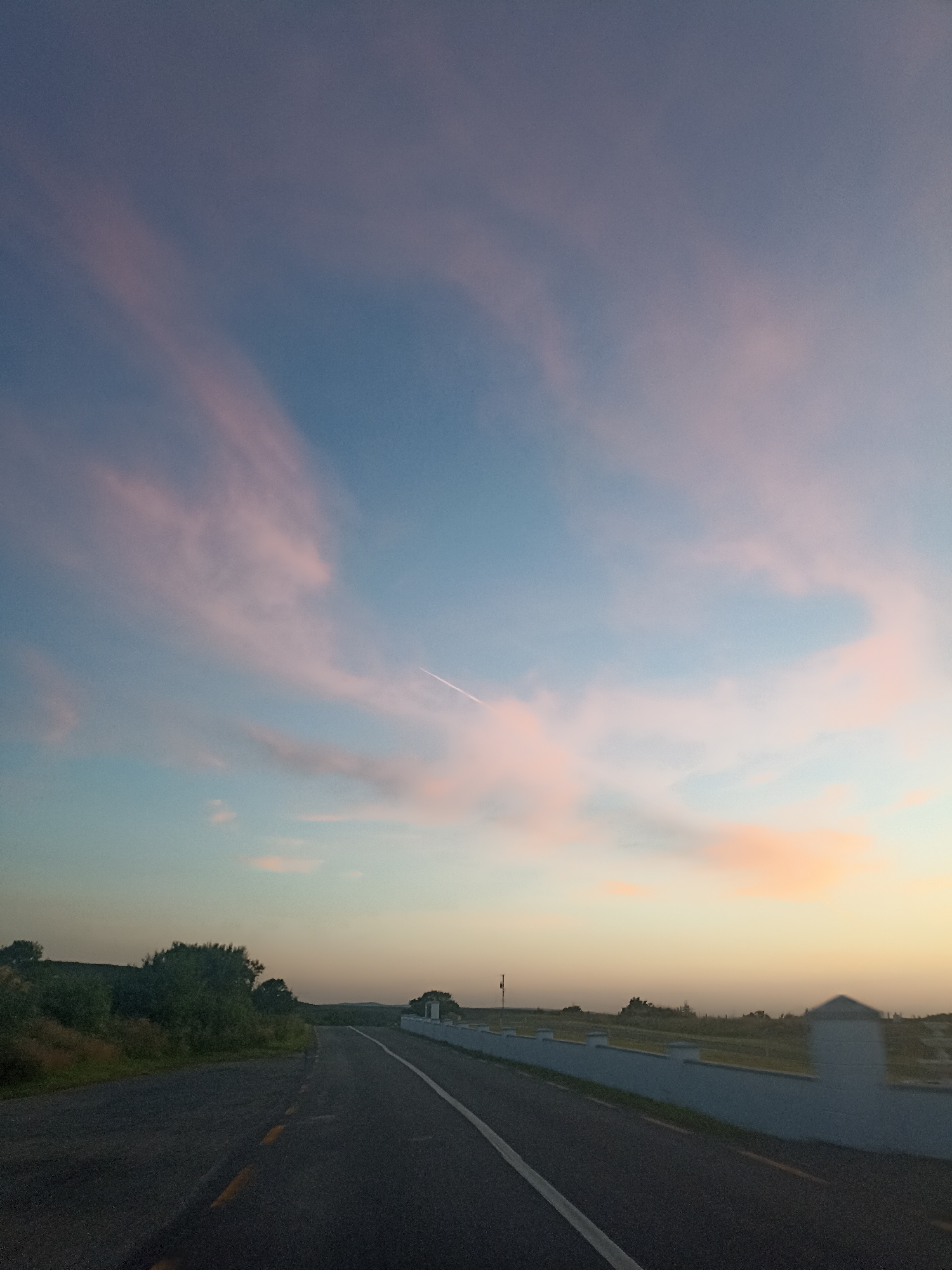 This is a vertical, eye-level, medium shot of a road at dusk in Ranafast, Ireland. The sky is a gradient of blue and pink with wispy clouds and a contrail. On the left, a grassy hillside is covered in trees and shrubs. On the right, a white concrete wall with decorative pillars runs alongside the road. The road itself is asphalt with a white line down the center and yellow dashed lines on the shoulder. The environment is rural and serene.