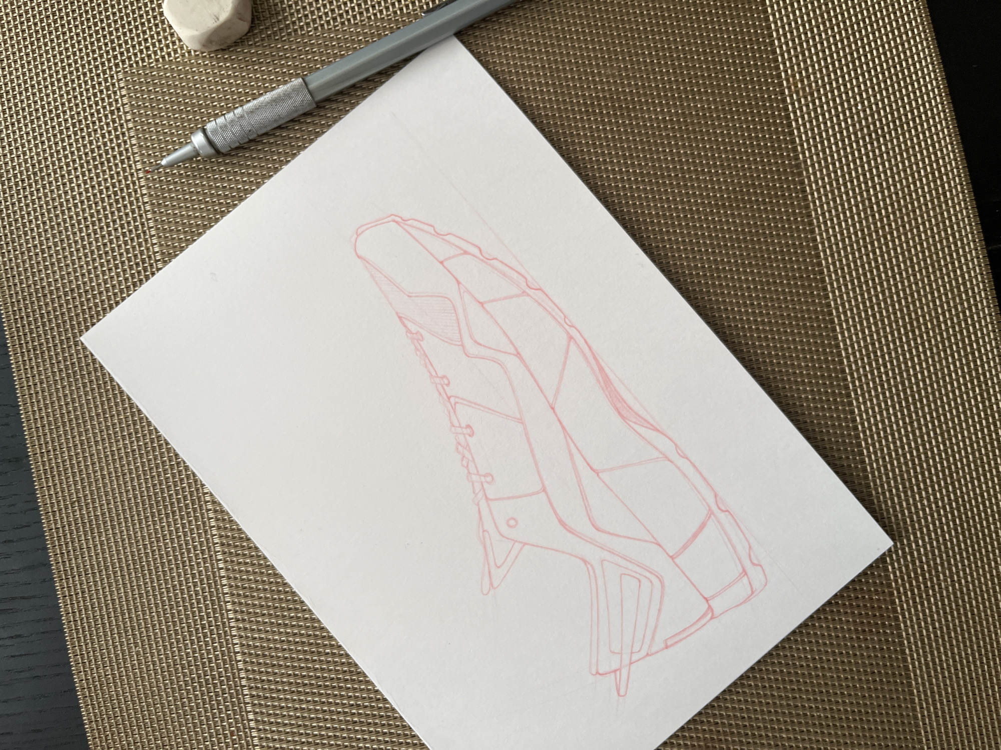 On a textured brown placemat sits a white piece of paper with a red line drawing of a complex object, possibly a piece of machinery or vehicle component. A mechanical pencil and a white eraser are located at the upper left corner of the image. The drawing occupies the center of the paper, oriented diagonally. To the lower left and right edge of the placemat a dark wooden surface is visible. The location is Unknown, Unknown.