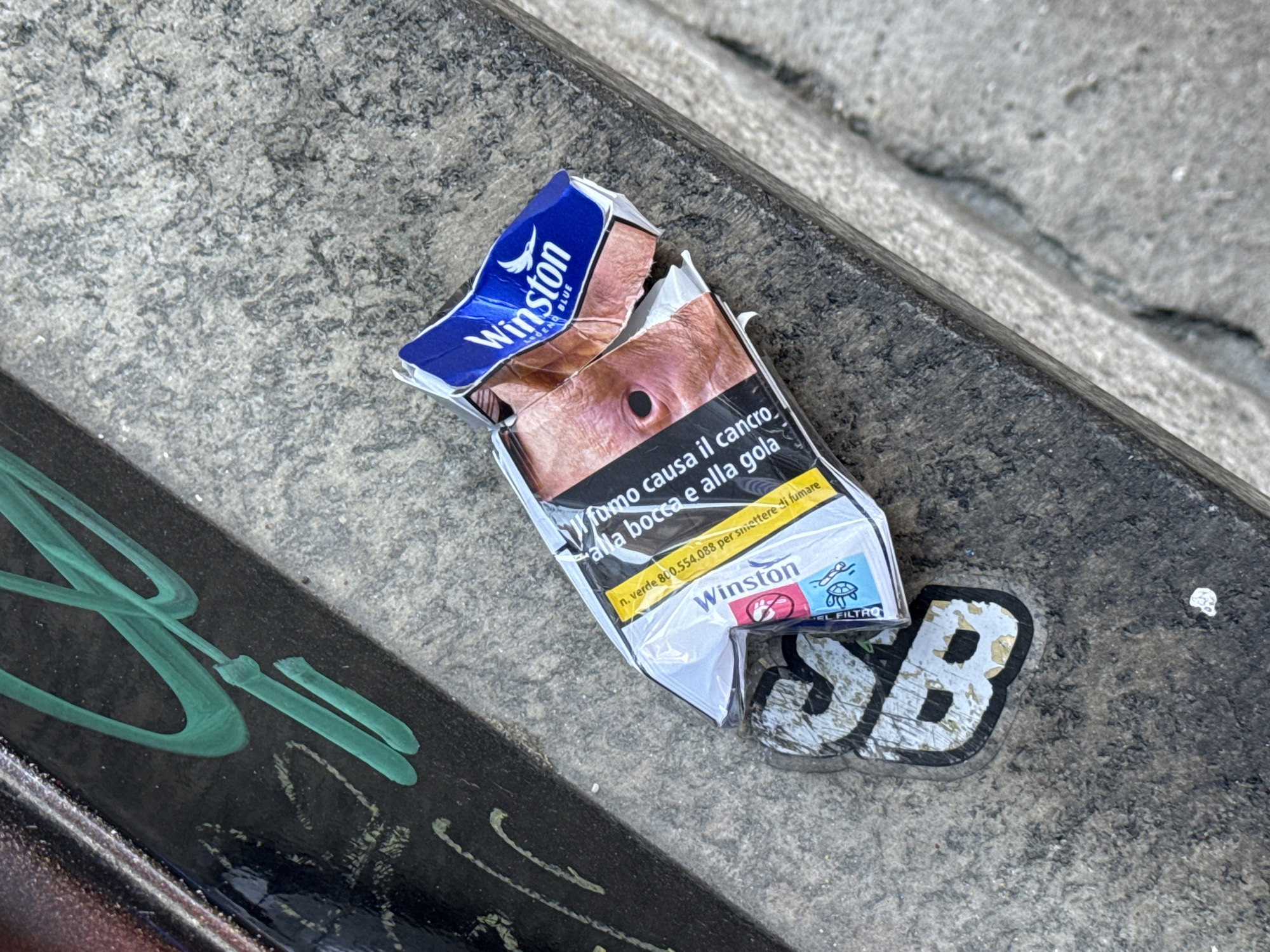 The image shows a discarded and crushed Winston cigarette pack lying on a weathered, grey surface, possibly concrete or stone. The pack is torn open, revealing a graphic image of a person's face damaged by cancer, along with the Italian warning "Il fumo causa il cancro alla bocca e alla gola" ("Smoking causes cancer of the mouth and throat"). The Winston brand logo is visible, along with a phone number for quitting smoking. Next to the cigarette pack is a worn sticker with the letters "SB" in a stylized font. A green, graffiti-like marking is also visible in the lower-left corner of the frame. Based on the Italian language on the pack and the urban setting, the location is likely Milan, Italy.