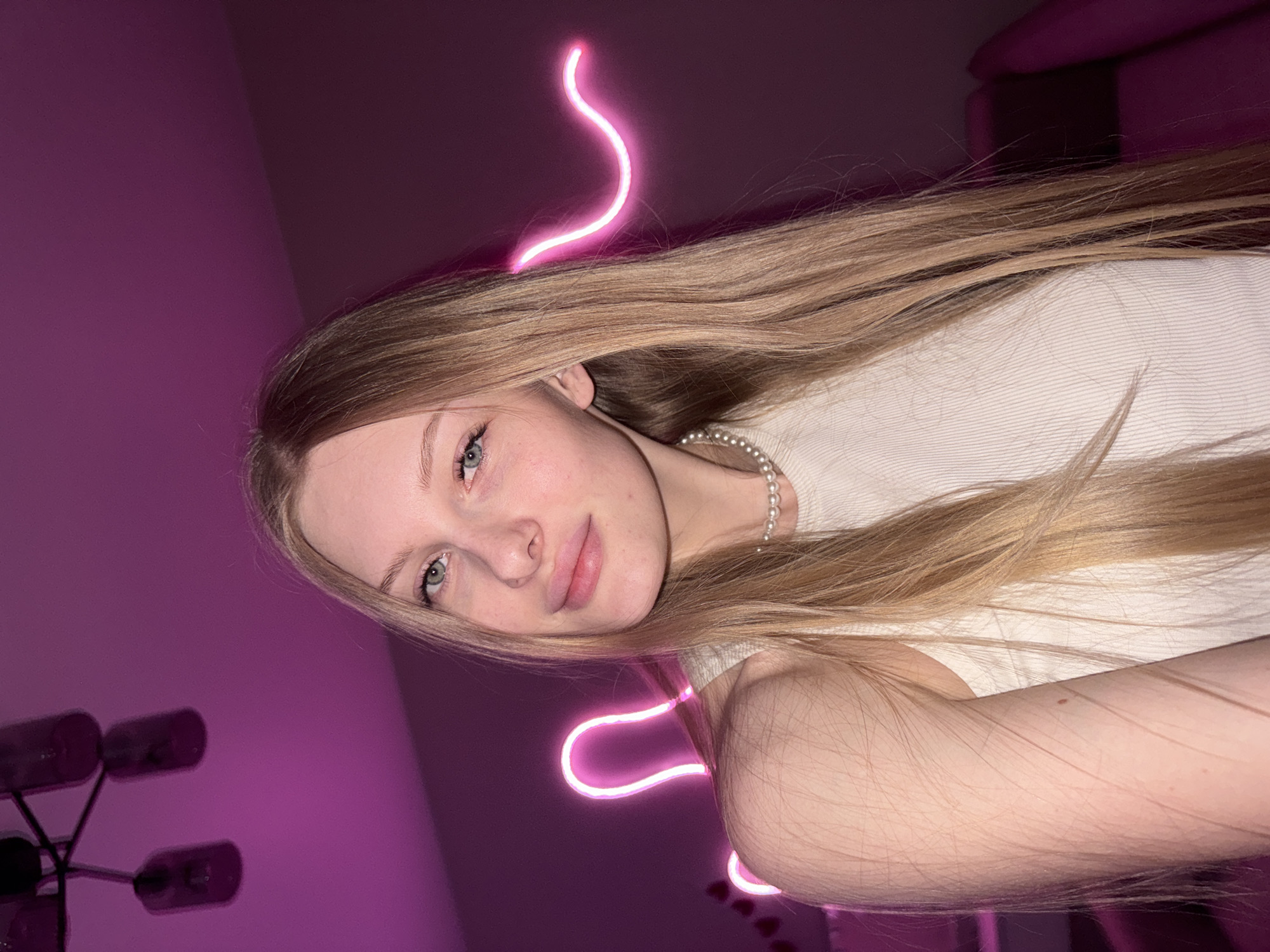 A portrait of a young woman is captured at an unconventional, rotated angle. She appears to be in her late teens or early twenties, with long, straight, light brown or blonde hair flowing down her chest and arm. Her face is turned slightly towards the viewer, revealing light-colored eyes (likely blue or green) and a calm, soft expression with slightly parted lips. She wears a simple, light-colored (off-white or cream) ribbed top and a pearl necklace.
The background is predominantly a deep magenta or purple, suggesting a room with distinct artificial lighting. A bright, wavy pink neon light is a prominent feature, curving across the top and bottom sections of the frame, enhancing the modern, vibrant ambiance. In the lower left corner, a dark, multi-bulb light fixture, resembling a chandelier, is partially visible against the purple wall. The setting is indoors, likely in the evening or at night due to the reliance on artificial light. The image conveys a serene and stylish mood, indicative of a posed photograph, possibly a selfie. No text is visible. The location context is Perm, Russia.