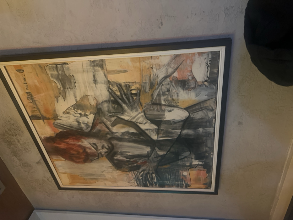 A vertically oriented, rectangular framed painting is affixed to a textured light grey wall within a building in Smederevska Palanka Municipality, Serbia. The artwork is an abstract-figurative piece depicting a human figure, likely female, rendered predominantly in shades of grey, black, and white, with prominent red hair styled in an updo. The figure's head is tilted downward, and both hands are dynamically positioned; the left hand is raised near the figure's chest or face, while the right hand is extended to the right, fingers splayed. The background of the painting features broad, abstract brushstrokes in warm tones of orange, beige, and darker grey, suggesting a dynamic, layered composition. A discernible artist's signature and the year "2022" are visible in the lower right corner of the painting. The artwork is enclosed within a frame featuring a thick dark outer border and a thinner white inner border. The wall itself exhibits a rough, stucco-like texture. In the upper left corner of the scene, a brown architectural element, possibly part of a ceiling or beam, is partially visible. The lower right foreground includes a partial view of a dark, soft object, consistent with a cushion or furniture armrest.
