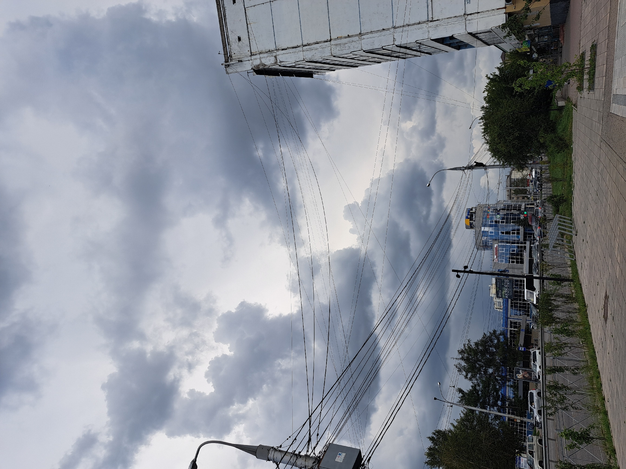 The image depicts a cloudy sky over a city street scene in Irkutsk, Russia. The foreground shows a paved sidewalk with a metal fence and some greenery. A street is visible behind the fence, with several cars and trucks driving. On the left side of the street, there are trees and buildings, including a blue glass-fronted building with a sign. On the right side of the street, a white multi-story building with windows is prominent, along with a street lamp and an abundance of overhead power lines. The overall impression is of a typical urban environment under overcast skies.