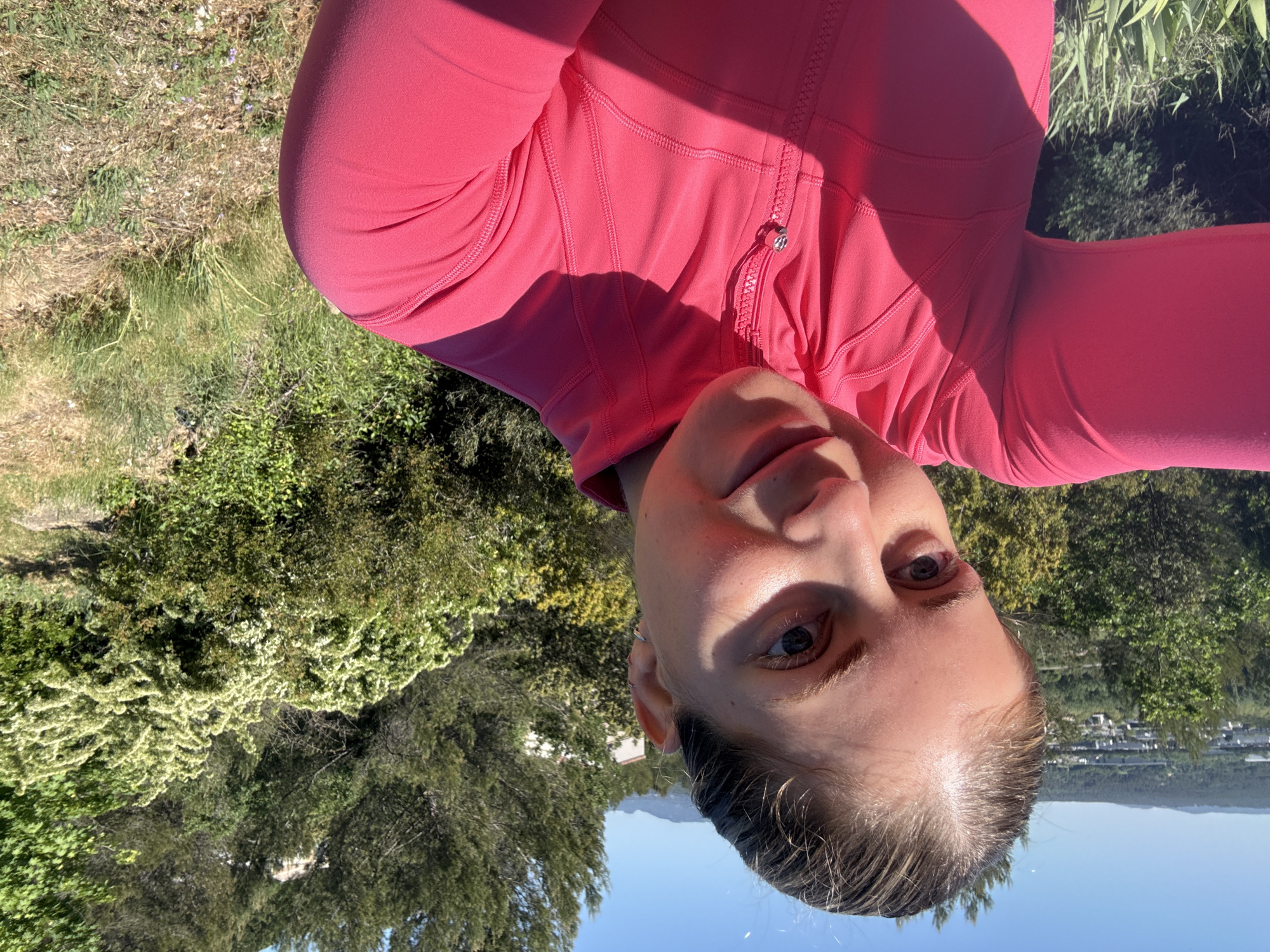 A medium-shot photograph captures a woman in a bright pink zip-up jacket. She is positioned outdoors against a backdrop of greenery. Buildings and a mountain range are visible in the background, with a patch of blue sky. The location is Rousset, France.