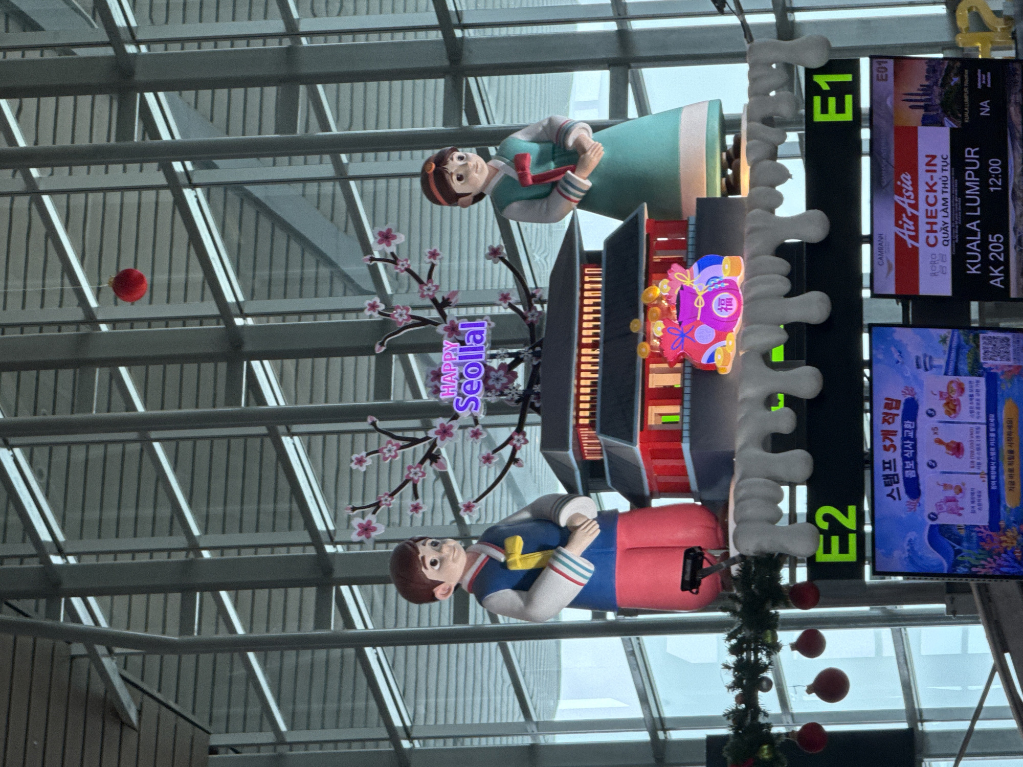 The image captures a festive display at Cam Ranh International Airport in Bắc Cam Ranh, Vietnam, likely for a Lunar New Year celebration. In the foreground, two cartoon figures dressed in traditional Korean Hanbok stand on either side of a miniature Korean palace structure. The male figure is on the left, wearing a blue outer garment over a white shirt and red trousers. The female figure on the right is in a teal Hanbok. Above them, a cherry blossom branch with pink flowers extends across the frame, and a red spherical ornament hangs from the ceiling. The palace features red walls and a dark roof, with illuminated windows. Below the display, a digital sign shows "E2" and another shows "E1", indicating departure gates. Further down, a screen displays flight information for AirAsia, with a destination of Kuala Lumpur. The overall environment is that of a modern airport terminal with a high ceiling and metallic supports.