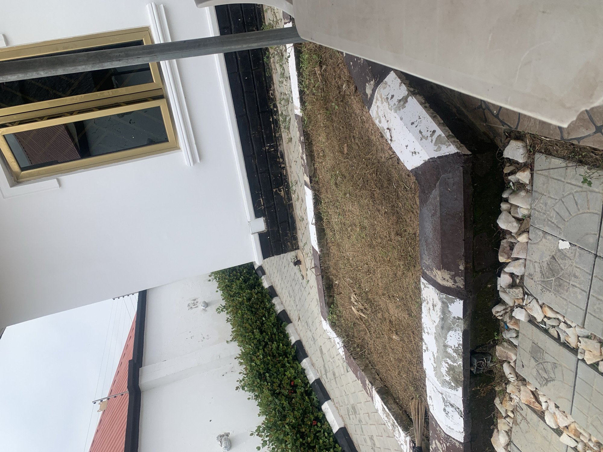The image depicts a section of an outdoor area, likely a courtyard or garden, adjacent to a modern white building with gold-trimmed windows. The ground is paved with light gray interlocking stones, interspersed with scattered light-colored rocks. A dried, grassy area is bordered by a low concrete structure painted with a black and white striped pattern. To the left, a dense green hedge runs along a white wall, which has a red-tiled roof and an electric fence visible at the top. A dark object, possibly a bird, is perched on the paved area near the hedge. The overall scene suggests a residential or commercial property in Jalingo, Nigeria, characterized by its construction style and landscaping elements.