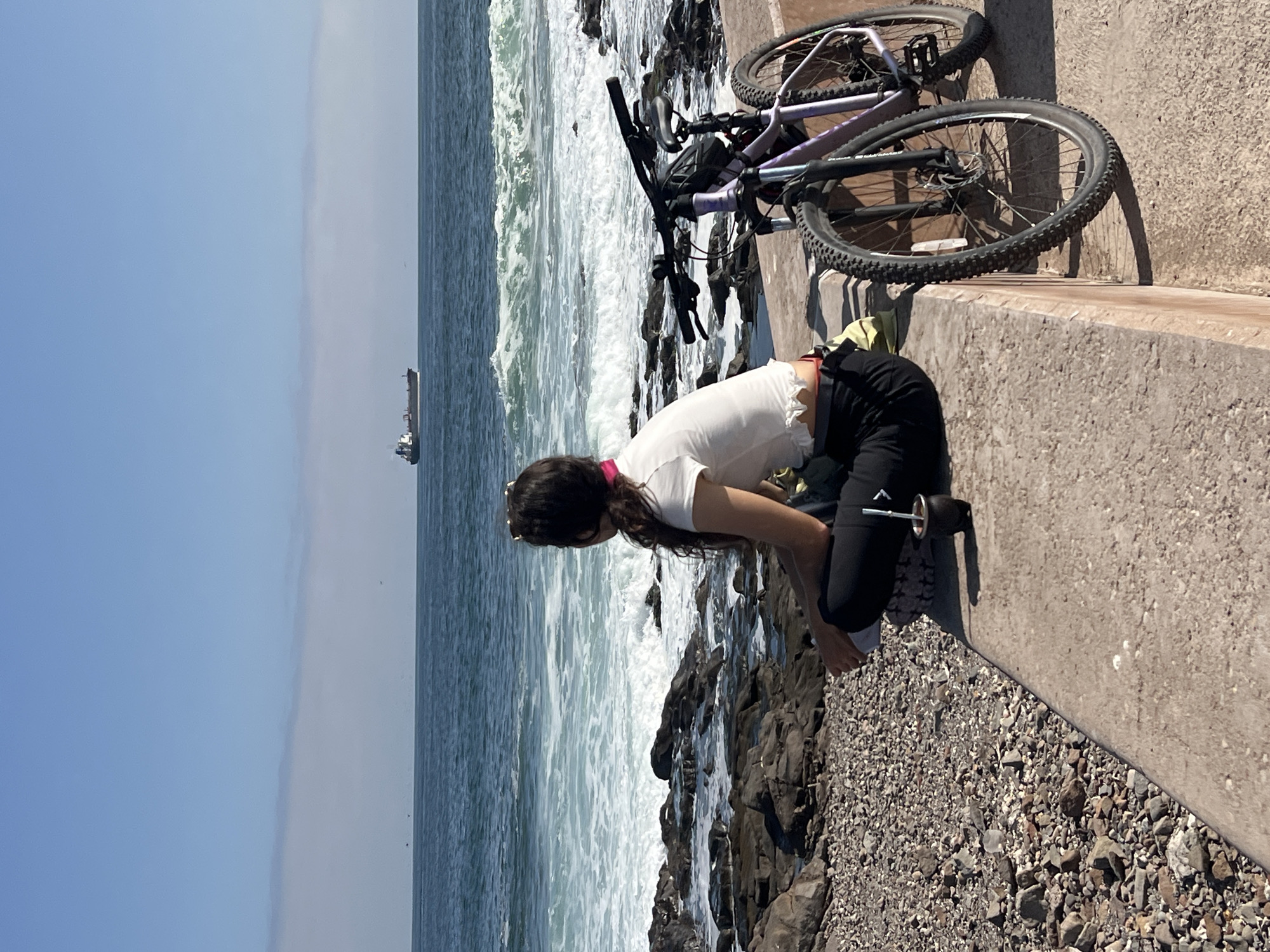Arica, Chile: A woman bends at the waist toward a rocky beach next to the ocean, with a ship visible in the distance. She is wearing a white t-shirt and black leggings. A bicycle rests on a low concrete wall adjacent to the beach. The sea has white-crested waves.