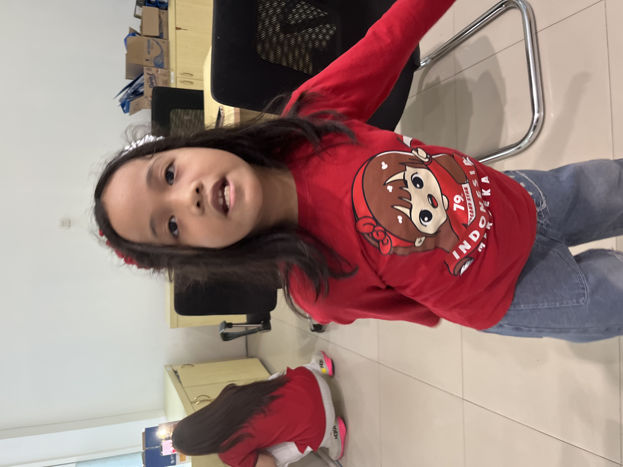 The image shows two young girls wearing red shirts. The girl in the foreground is looking at the camera, with her mouth slightly open. She has long, dark hair and is wearing a red shirt with a cartoon character and text that appears to say "INDONESIA". She is also wearing denim jeans. To the left, another girl is crouching down, facing away from the camera. She is also wearing a red shirt and white pants, with brightly colored sneakers. The background appears to be an indoor space, possibly an office or classroom, with light-colored walls, light wood cabinets, and black chairs. There are some cardboard boxes on top of the cabinets. The floor is tiled with large, light-colored square tiles. The overall lighting is bright and even, suggesting an indoor environment.