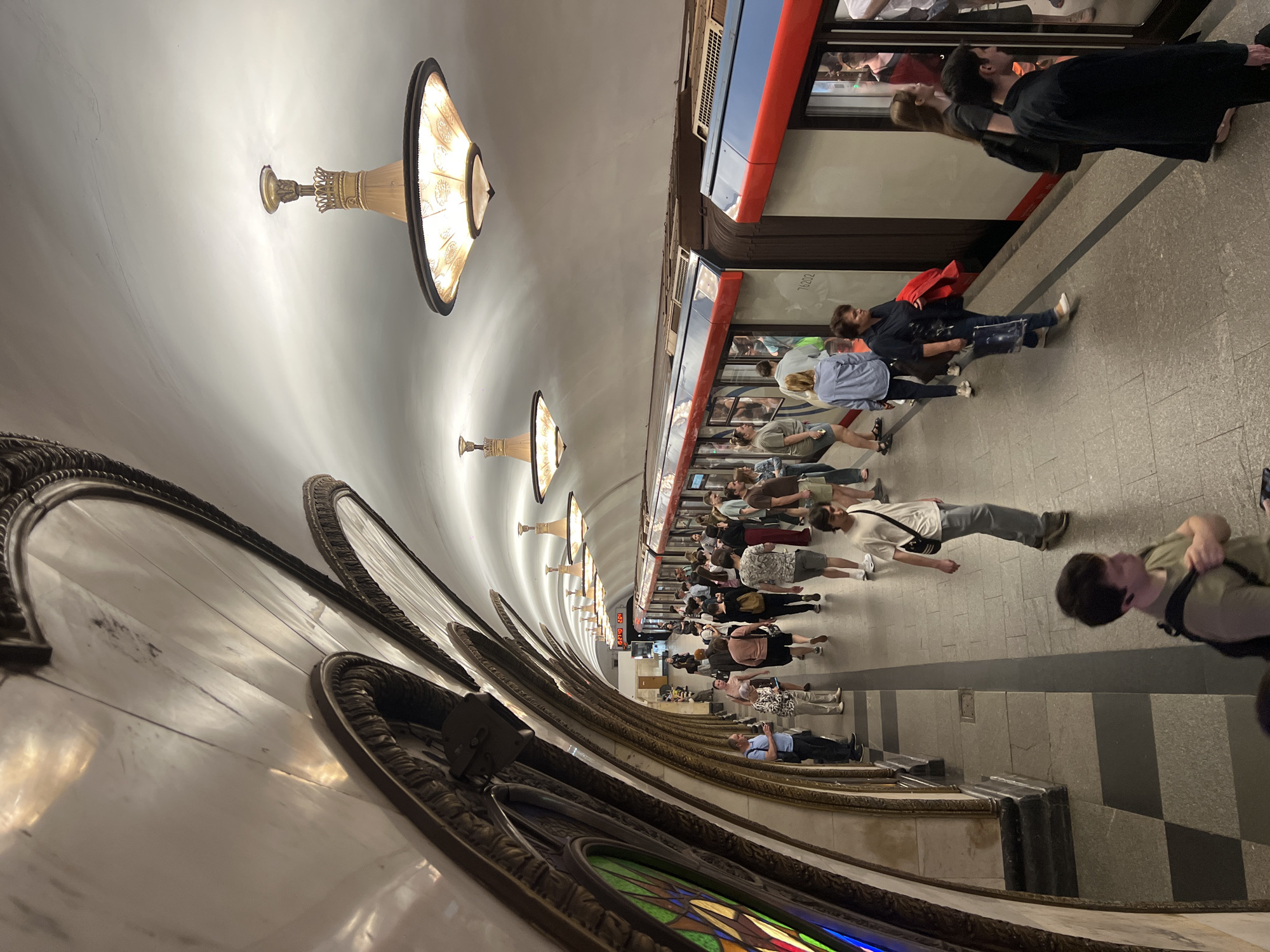 The image captures a scene inside a Moscow metro station, Russia. A long row of people is standing along the edge of the platform, waiting for a train. The train is stopped at the platform, the doors are open, and passengers inside the train are visible. Overhead, decorative light fixtures line the ceiling of the station. A large, ornate architectural element runs along the wall on the left side of the image, with stained glass visible on its lower portion. The floor is tiled.