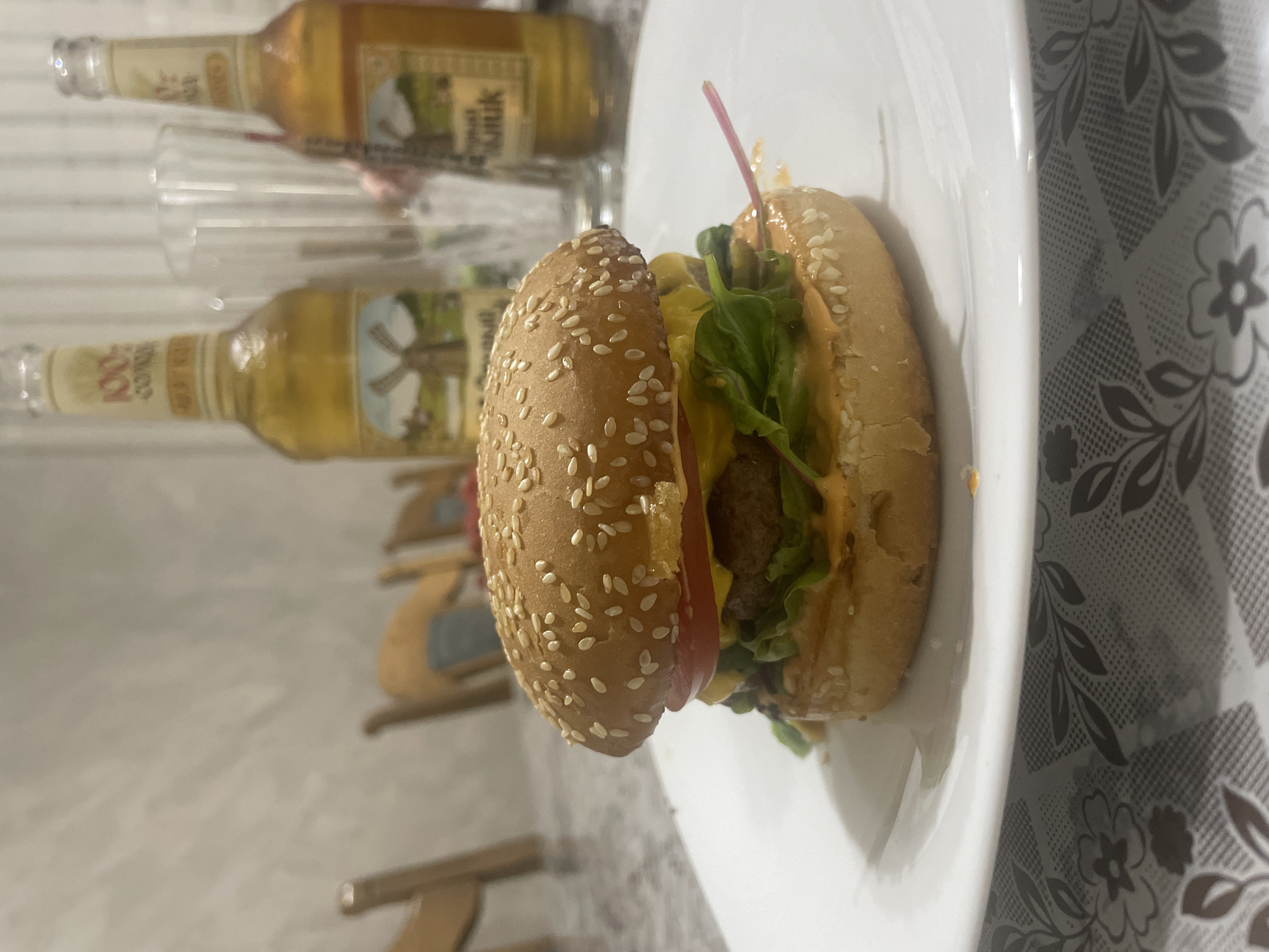 The image depicts a close-up view of a gourmet hamburger resting on a white plate. The hamburger is characterized by a sesame seed bun, a thick patty, melted cheese, a slice of tomato, and fresh lettuce. In the background, two bottles of beer and a clear glass are visible, suggesting a casual dining setting. The lighting is soft, creating a warm and inviting atmosphere. The scene is set indoors, likely in a home environment, with a table covered by a patterned tablecloth. The overall composition emphasizes the appetizing presentation of the food.