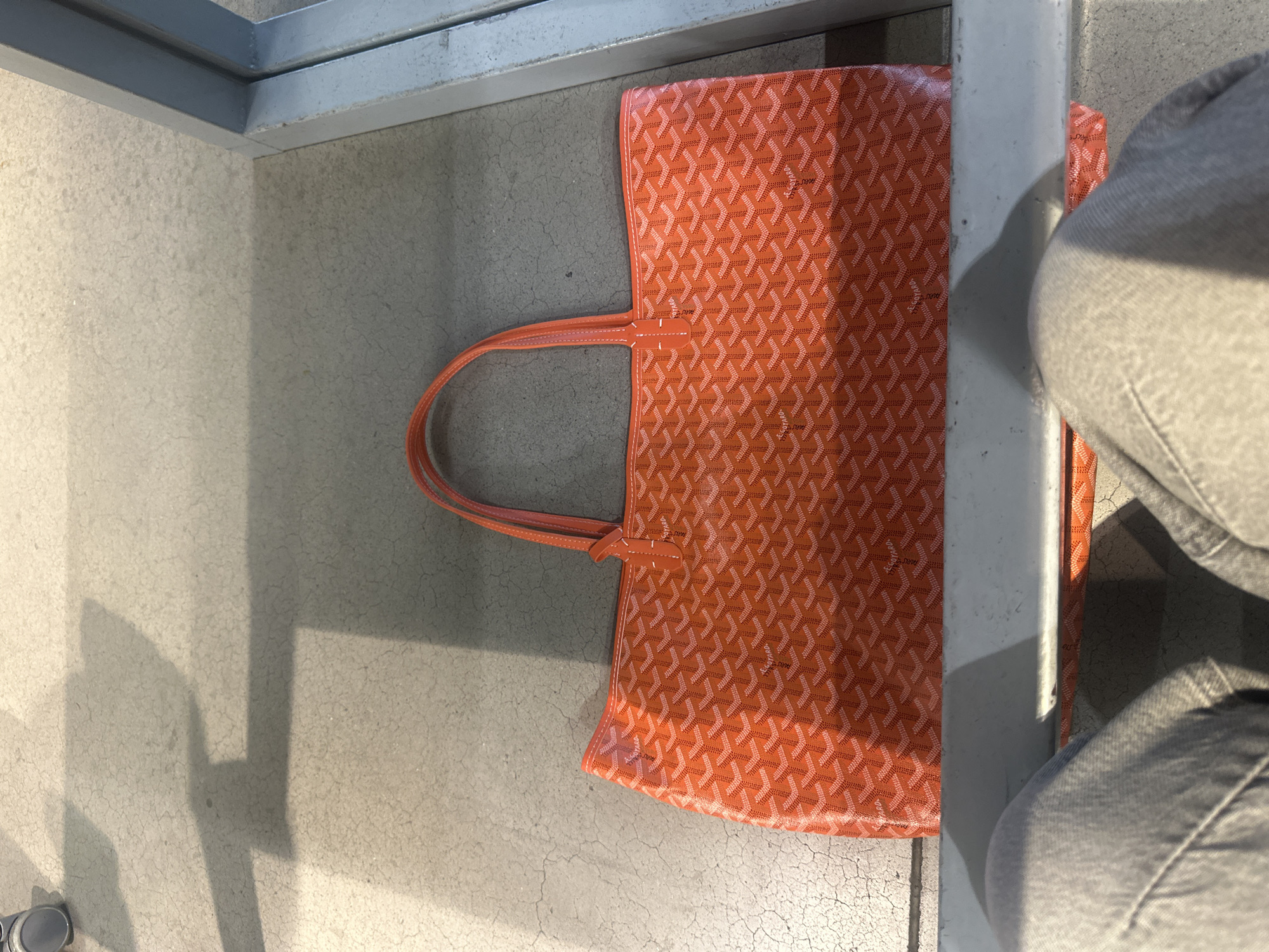 The image depicts an orange Goyard tote bag placed on a concrete surface, likely a floor, with metal frames surrounding it. A pair of jeans are partially visible on the right side of the frame. The bag features the signature Goyardine pattern. The environment appears to be a neutral, indoor space. No information in the picture explicitly indicates it's in Mexico City.