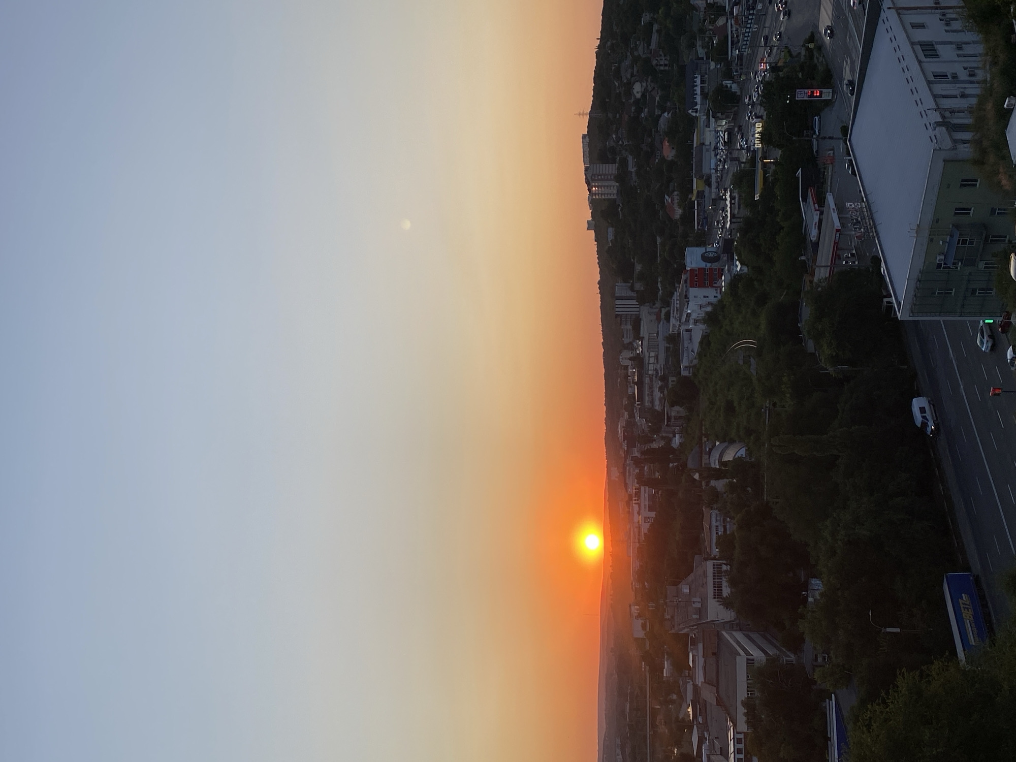 This is an aerial, high-angle, vertical shot of a city at sunset. The sun is a bright orange orb low on the horizon, casting a warm glow across the sky and illuminating the cityscape. The sky transitions from a pale yellow near the horizon to a soft blue at the top of the frame.
The foreground is dominated by buildings, some with flat roofs and others with more complex architectural features. Trees are interspersed throughout the urban landscape, some forming dense clusters and others lining streets. A highway runs through the scene, with several vehicles visible, including cars, a van, and a truck with a blue trailer. A gas station with visible signage is also present. In the background, rolling hills are covered in greenery, with buildings scattered on their slopes.
The overall impression is one of a developing urban area bathed in the soft, fading light of sunset. The scene is devoid of any visible people.
Based on the presence of the "GRAND" signage and the architectural style of the buildings, this location is likely within the city of Chișinău, Moldova.