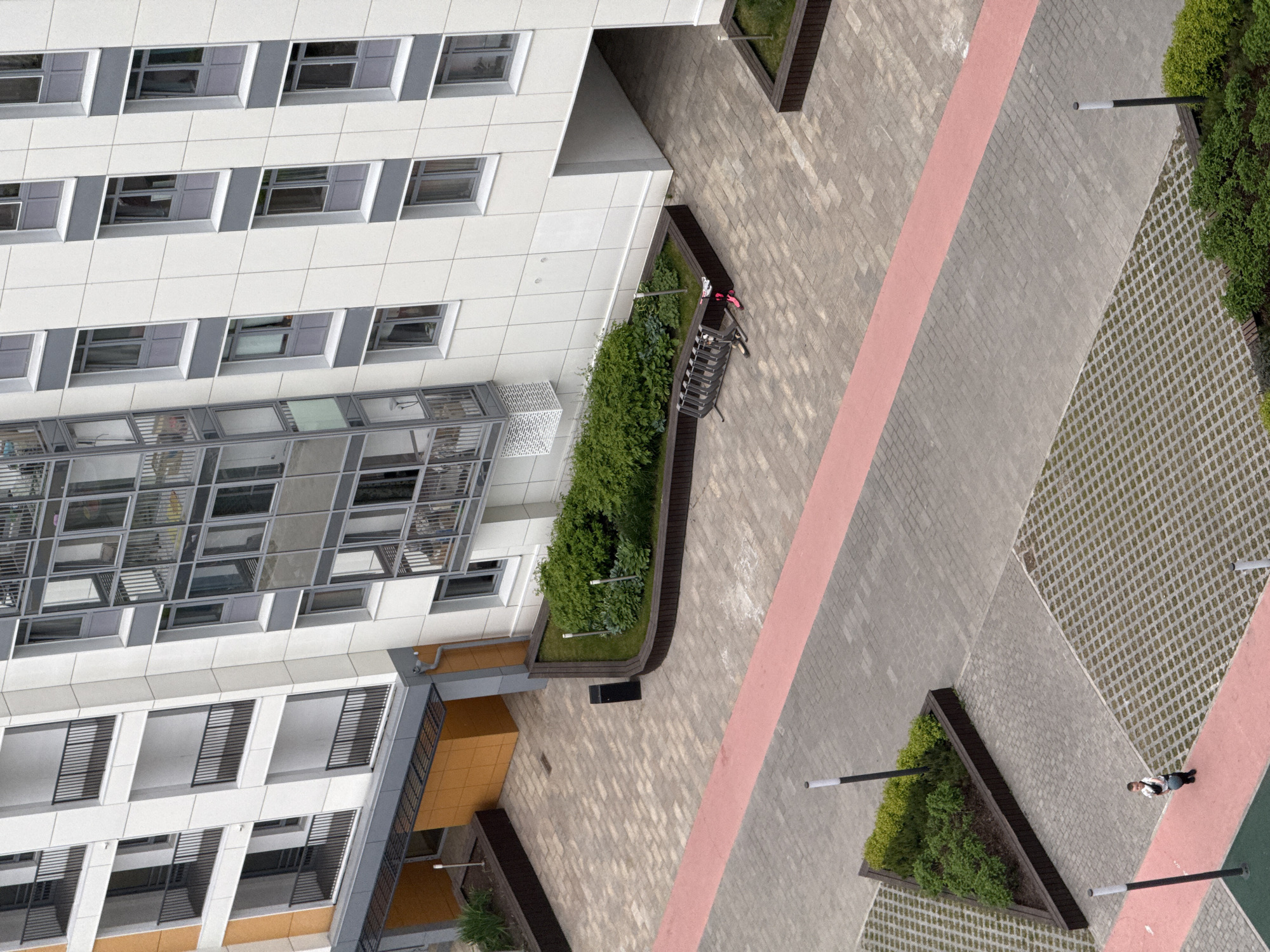 The image shows an aerial view of a building and its surroundings in Perm, Russia. The building has a modern design with white and gray facade elements and numerous windows. A pedestrian area with patterned brickwork, pink stripes, and grass-filled concrete grids is adjacent to the building. There are planters with greenery and lamp posts along the walkway. A person is standing in the lower right corner of the image. A bicycle is parked next to the building.