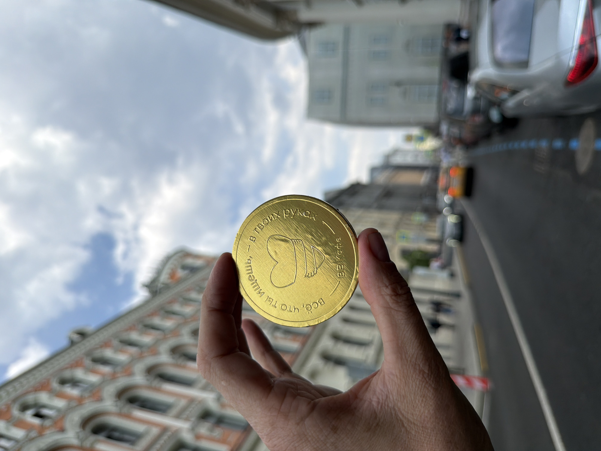 A hand holds a golden coin with Russian inscriptions in front of a street scene in Moscow, Russia. The coin features an icon of a heart with legs and the text "В ТВОИХ РУКАХ, - всё, что ты ищешь, BB.Кафе" written around the perimeter. The background shows a blurred cityscape with ornate buildings, cars, and a cloudy sky, indicating an urban environment. The buildings exhibit distinct architectural styles, and the street appears active with pedestrian and vehicular traffic. The photograph's perspective is angled upwards, focusing on the coin as the primary subject against the backdrop of the city.