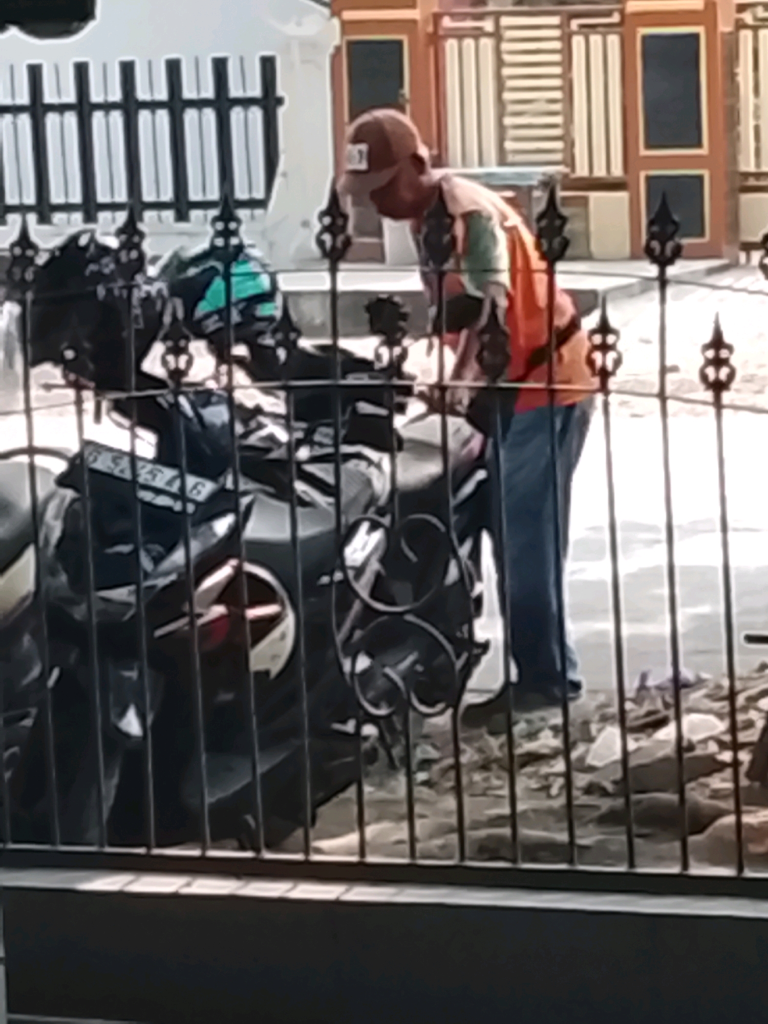 A person wearing a baseball cap and a colorful shirt over blue jeans is seen tending to a motorcycle parked behind a black metal fence. The motorcycle is dark-colored, with a visible license plate. The background shows a building with patterned doors and a fence, suggesting an urban or suburban setting in Slawi, Indonesia. The scene is captured in daylight.