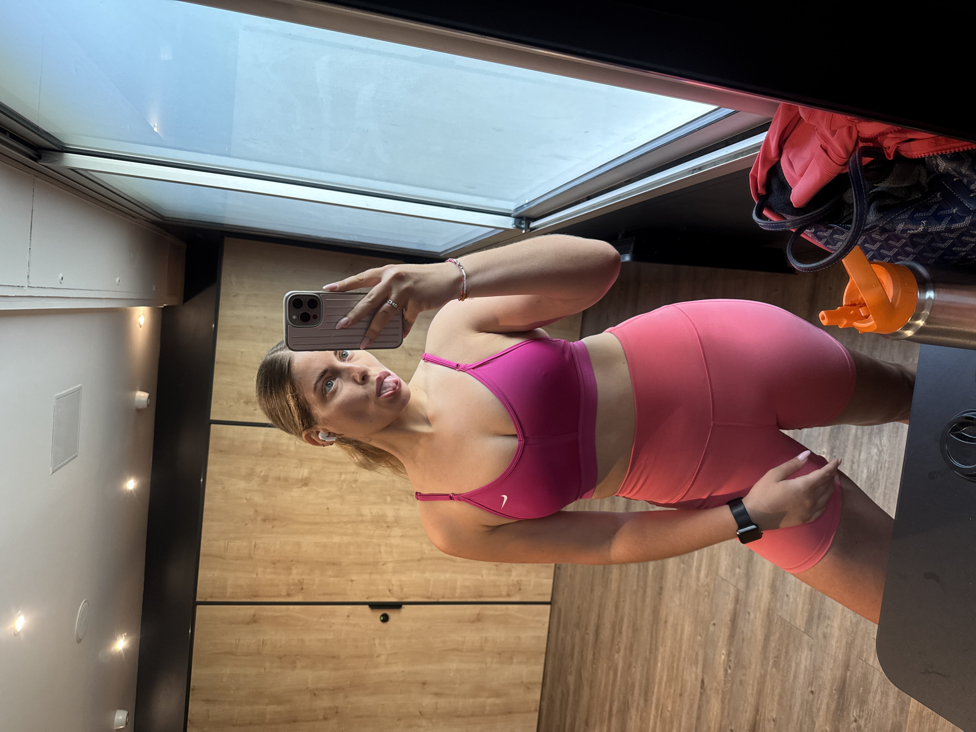 A woman stands in a room with wood-paneled walls and a large window, taking a mirror selfie. She is wearing a pink sports bra and pink biker shorts, with white earbuds in her ears. Her tongue is sticking out, and she is holding her phone in her right hand. A water bottle and a gym bag are visible in the foreground. The room appears to be a gym or locker room.