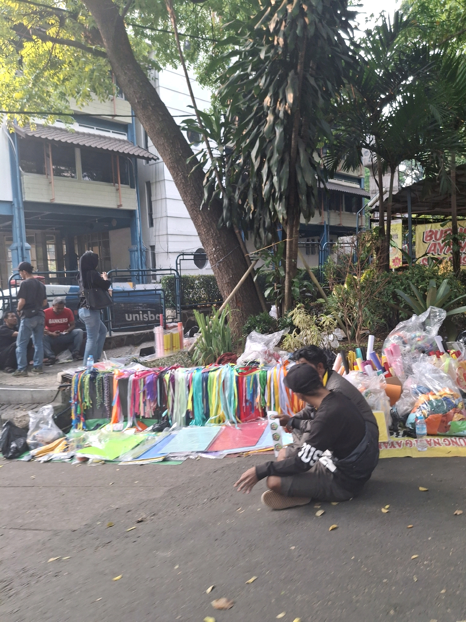 The image captures a street scene in Bandung City, Indonesia, where several individuals are gathered around a makeshift stall displaying various colorful items. The stall, set up on the asphalt ground next to a sidewalk, features a collection of brightly colored ribbons, fabric samples, rolls of colored paper, and wrapped goods. Two men are seated on the ground near the stall, appearing to be engaged with the merchandise. One man, wearing a black long-sleeved shirt with white lettering, is looking down, possibly examining an item. The other man, wearing a grey long-sleeved shirt, is partially visible. In the background, a building with multiple stories and a covered walkway can be seen, along with trees and lush greenery, suggesting an urban setting with some natural elements. Several other people are visible in the background, some standing and some sitting, near a blue fence with "unisba" printed on it. The overall atmosphere suggests a lively outdoor market or a street vendor operation.