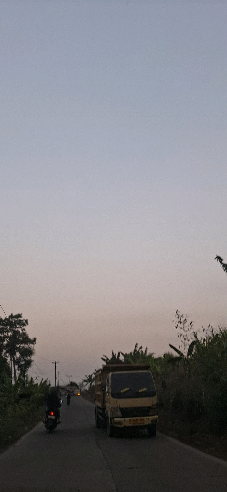 The image depicts a road scene at dusk in Cibinong Hilir, Indonesia. The sky is a gradient of pale blue to pink, suggesting twilight. A yellow dump truck is driving on the right side of the paved road. To the left of the truck, a motorcycle with a rider wearing dark clothing is moving away from the camera. Further down the road, another motorcycle is visible. Lush green vegetation, including banana trees, lines both sides of the road. Electrical poles and wires are also present. The overall atmosphere is calm and rural.