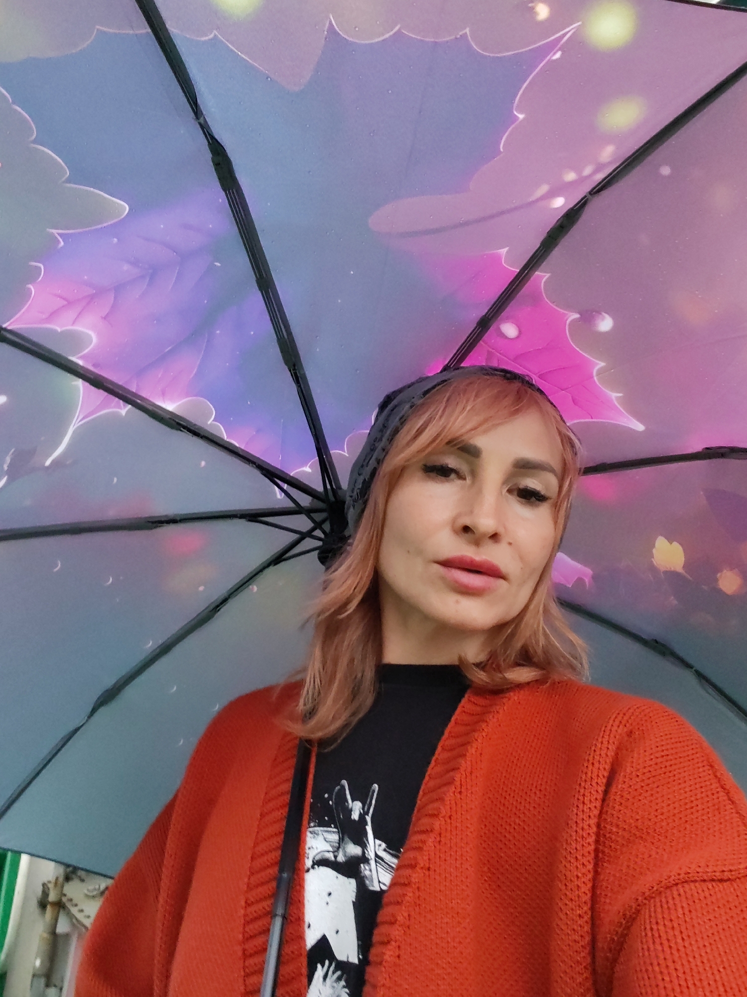 In this selfie, a woman with light pink hair, wearing a dark grey beanie and an orange cardigan over a black t-shirt, is looking at the camera. She is holding an umbrella over her head, which has a colorful pattern of leaves and glowing orbs in shades of purple, pink, and blue. The umbrella's spokes are visible, dividing the frame.
The woman has makeup on, with defined eyebrows and lipstick. Her expression is neutral. The lighting suggests it might be daytime, possibly overcast or rainy, given the umbrella. There are water droplets visible on the umbrella's fabric, indicating wet weather. The background is out of focus but suggests an outdoor urban environment with some metal structures visible on the left.
The scene is likely set in Aleksandrovskoe, Russia, as indicated by the location context. No visible text is present in the image.