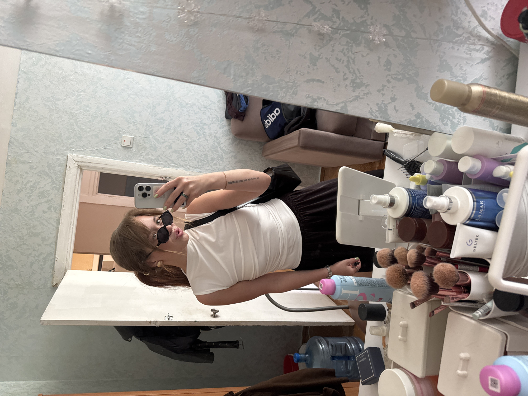 A selfie taken in a mirror. The subject is a fair-skinned woman with brown hair wearing a white t-shirt and dark bottoms. She's also wearing sunglasses and holding a smartphone to take the picture. The mirror reflects a room with patterned wallpaper and furniture. In front of the mirror, there is a vanity with makeup brushes, toiletries, and water bottles. Based on the presence of Russian language labels and the prompt, the location is Chaikovsky, Russia.