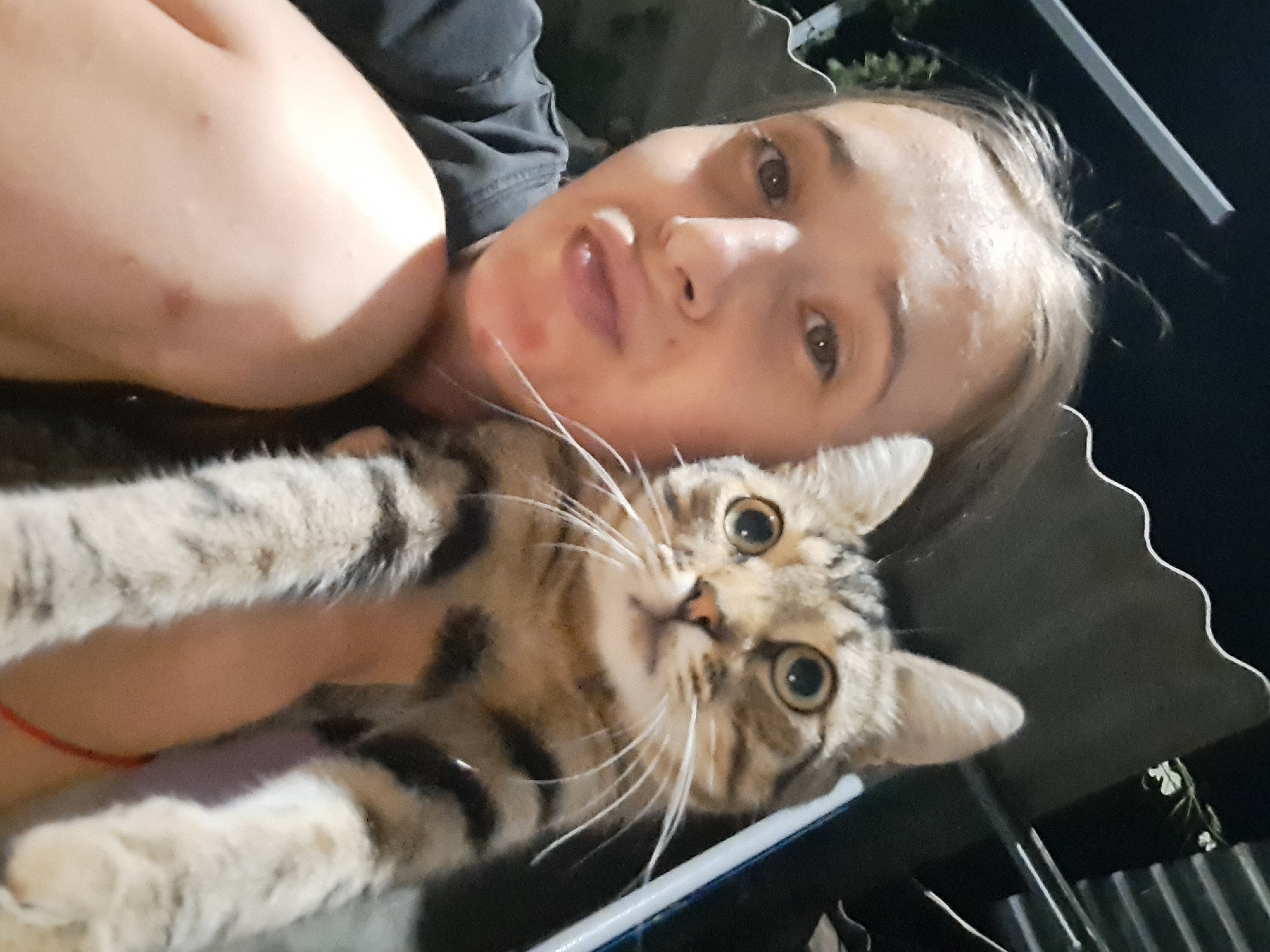 A close-up, selfie-style photo shows a young woman with light brown hair holding a tabby cat. The woman is looking at the camera with her mouth puckered. She is wearing a dark t-shirt. The cat, held in her arms, is looking at the camera with wide eyes. The background is dark, suggesting it is nighttime. There are some corrugated metal structures and lights visible in the background. The image appears to have been taken outdoors, possibly on a balcony or patio. The lighting is artificial and somewhat dim. The location is stated to be Vypasne, Ukraine.