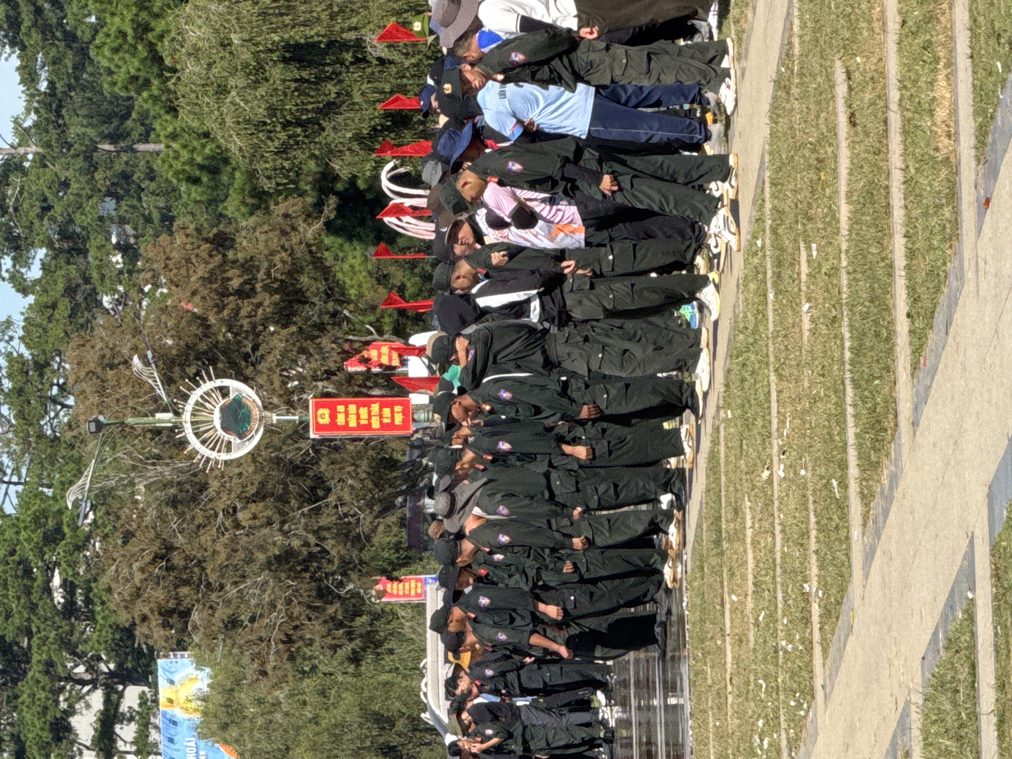 A group of people, mostly men, are standing in a line outdoors. They are wearing dark green uniforms with dark hats. Some are wearing caps, and others are wearing bucket hats. They are standing on a paved area that appears to be wet, possibly from recent rain or cleaning. In the background, there are trees and a tall white structure with decorative elements. Red flags are visible on poles. To the left, a large billboard with a picture on it is partially visible. The overall impression is that of an organized event or ceremony. The presence of Vietnamese writing on the flags and billboard suggests the location is Vietnam. Given the context of similar images and common gathering places in Da Lat, this scene is likely taking place at the Da Lat Flower Garden, a popular tourist attraction and event venue in the city.