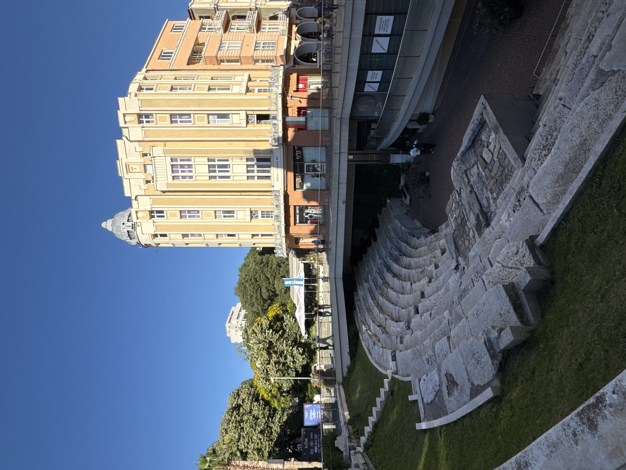 This is an eye-level, outdoor, full shot of the Ancient Roman Theatre in Plovdiv, Bulgaria, on a sunny day. The theatre, made of stone seating, is in the foreground. Behind it, a modern pedestrian walkway bridges over a lower level. A yellow and orange building with multiple windows, a dome, and some shops at its base, stands in the background. Trees and a clear blue sky complete the scene. Several people are visible walking on the walkway and around the theatre.