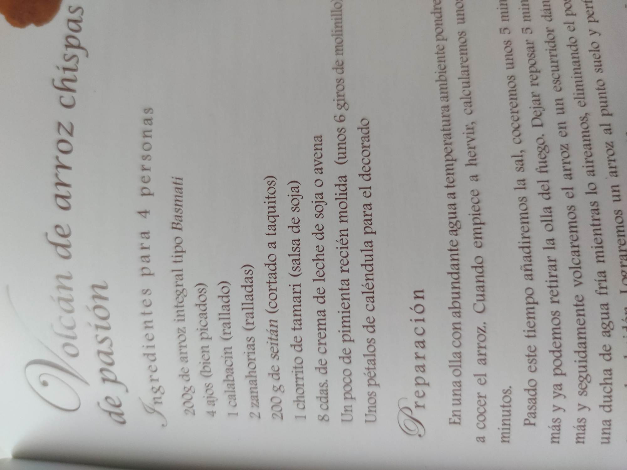 The image shows a page from a recipe book. The title of the recipe is "Volcán de arroz chispas de pasión" (Volcano of rice passion sparks). Below the title, it lists the ingredients for 4 people, including:
- 200g of Basmati whole grain rice
- 4 cloves of garlic (finely chopped)
- 1 zucchini (grated)
- 2 carrots (grated)
- 200g of seitan (cut into cubes)
- 1 dash of tamari (soy sauce)
- 8 tablespoons of soy or oat cream
- A pinch of freshly ground pepper (about 6 turns of the mill)
- Some calendula petals for decoration
Below the ingredients is the preparation section, which describes the steps to cook the rice.