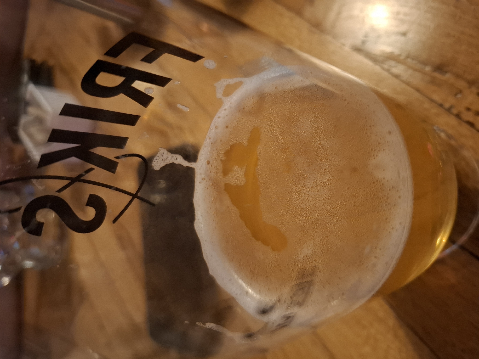 A close-up, angled shot shows the top of a glass filled with beer and its foamy head. The glass has the logo "FRIKS" etched into it, with the letters "S" and "F" partially visible, along with what appears to be a stylized "R" and "I". The beer is a golden amber color, and the foam is white and bubbly. The background is a wooden surface, likely a table, with some out-of-focus elements suggesting it's an indoor setting.
