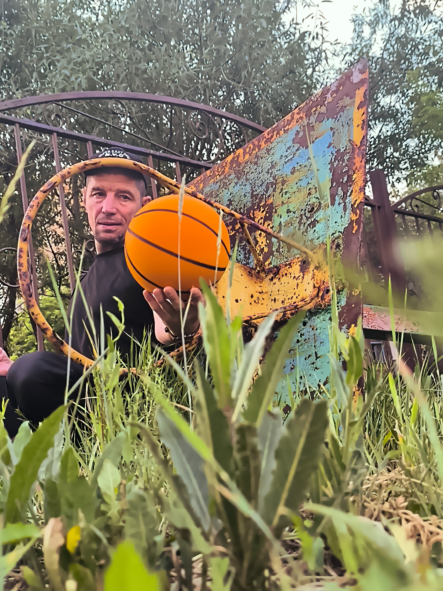 An adult male wearing a black t-shirt and a black baseball cap is pictured outdoors, holding an orange basketball. He is positioned behind a rusty, dilapidated metal structure with peeling paint in various colors, including yellow, blue, and brown. The man is partially obscured by tall green grass and foliage in the foreground. The background features trees and a dark metal fence. The scene appears to be in an overgrown, neglected area, possibly a park or abandoned lot. The image has a low-angle perspective, emphasizing the grass and the textured metal object.