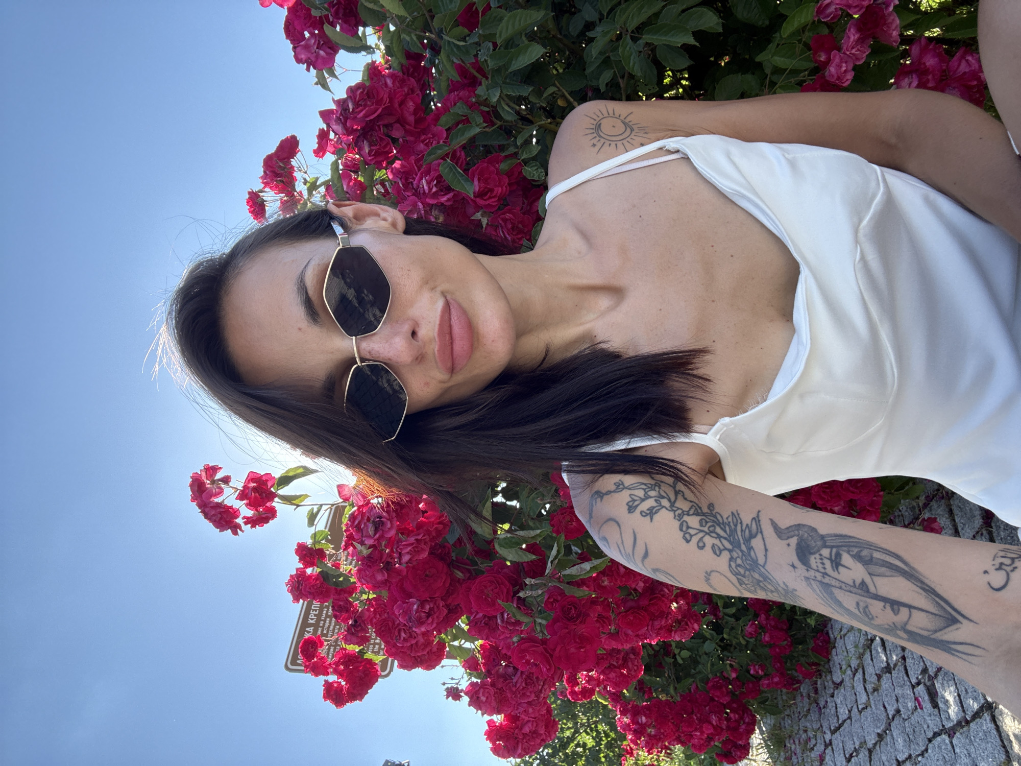 A medium shot captures a woman with long, dark hair and sunglasses, posing for a selfie in front of a vibrant pink rose bush. She is wearing a white slip dress and has tattoos on her left arm and shoulder. The background features a bright blue sky and a cobblestone path. A brown sign with text in Cyrillic is partially visible behind the rose bush. The overall scene suggests an outdoor setting, possibly in a park or garden, with the location identified as the Lovech Fortress in Lovech, Bulgaria.