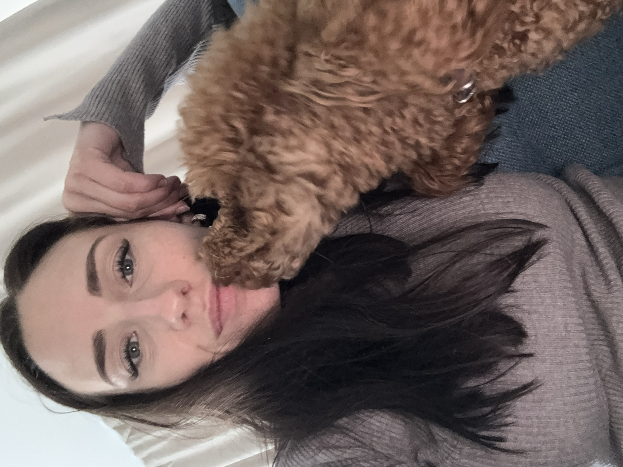 A light-skinned woman with dark hair and eyelashes lies on a white surface wearing a gray sweater. A reddish-brown poodle stands over her, possibly licking her face. The woman's right hand is raised near her ear. A blue textured surface is visible near the dog. The location is unknown.