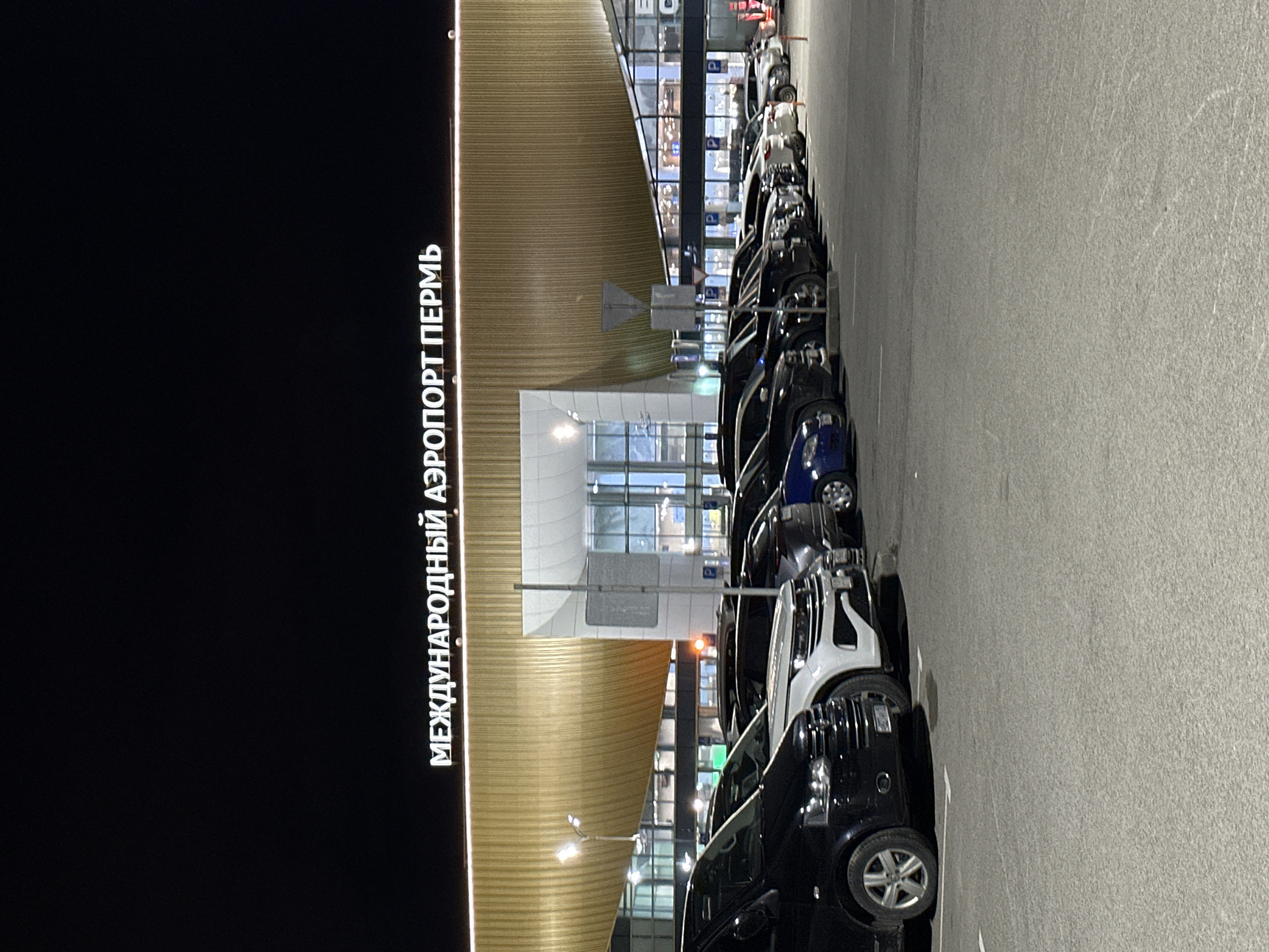 The image shows the exterior of the Perm International Airport in Bolshoye Savino, Russia at night. The airport building is partially visible, with the text "МЕЖДУНАРОДНЫЙ АЭРОПОРТ ПЕРМЬ" (International Airport Perm) displayed on a large sign. A line of cars is parked along the side of the airport facing toward the terminal.