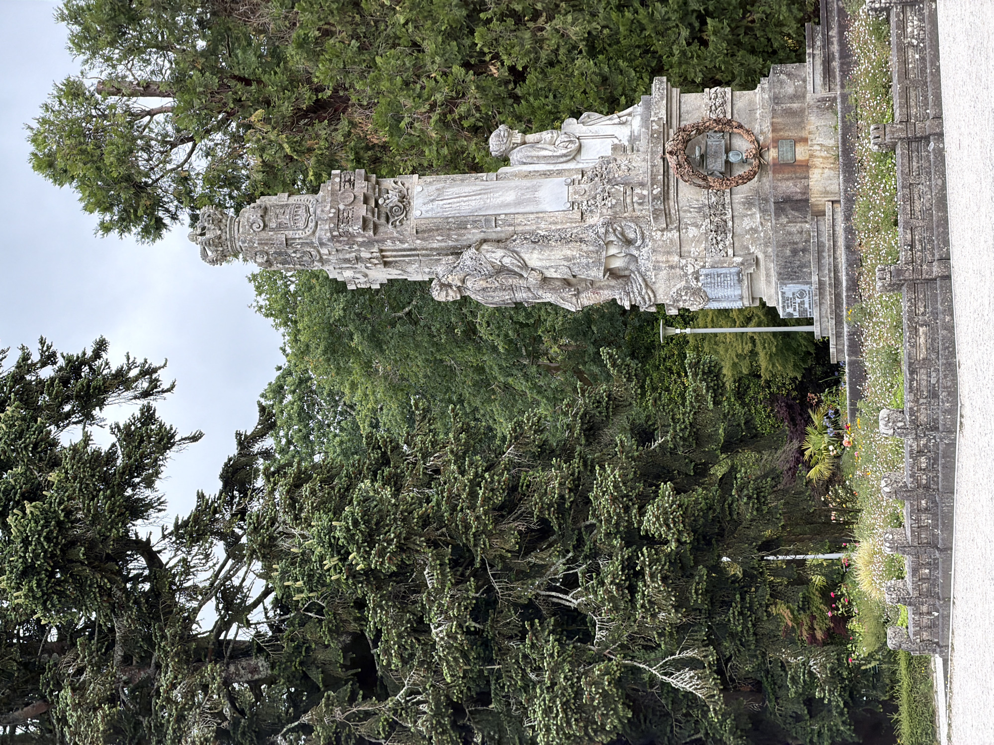 This is a vertical, eye-level shot of an ornate, weathered stone monument with a wreath and sculpted figures, set against a backdrop of lush green trees and a cloudy sky. The monument appears to be a memorial or a sculptural element within a park or garden. It features intricate carvings, including what looks like a seated figure at the top and other figures in relief on the main structure. A circular wreath, possibly made of dried branches or metal, is affixed to the monument. There are also plaques attached to the monument, though the text is not clearly legible. The surrounding vegetation is dense and varied, with evergreen trees dominating the scene. In the foreground to the right, a low wall or border is adorned with small white flowers. The overall impression is one of solemnity and permanence, hinting at a historical or commemorative purpose. The lighting suggests it might be daytime, possibly overcast.