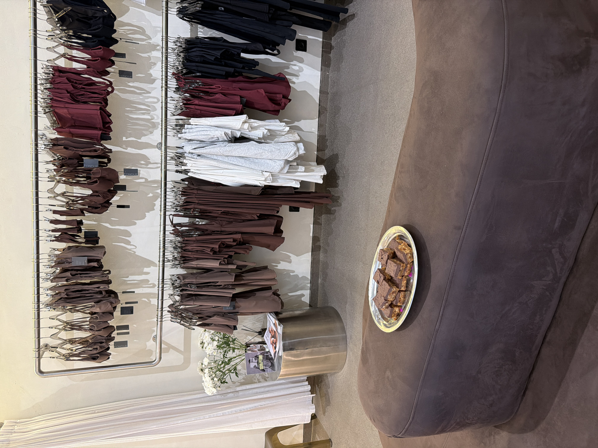 This is a medium shot of a retail space, possibly a boutique, with clothing items displayed on racks. The clothing appears to be women's activewear or loungewear, primarily in shades of brown, maroon, and black. On the left side of the frame, a white curtain hangs. In the middle ground, there is a silver cylindrical table with a vase of white flowers and some magazines. In the foreground, a brown upholstered bench or sofa is visible, with a silver platter of square-shaped, dark brown desserts placed on it. The floor is a neutral gray carpet. The lighting is soft and diffused, casting shadows from the clothing racks onto the wall. There are no people visible in the image. The scene is indoors, and the overall aesthetic suggests a clean, modern, and possibly upscale retail environment.