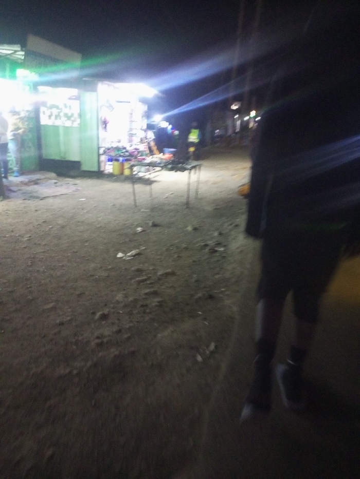 Night scene depicts a brightly lit vendor stall in the background. The vendor stall showcases an array of merchandise. Dirt ground leads to the stall. A person in dark clothing walks in the foreground of the image. Additional people can be seen near the vendor stall. Street lights illuminate parts of the background.