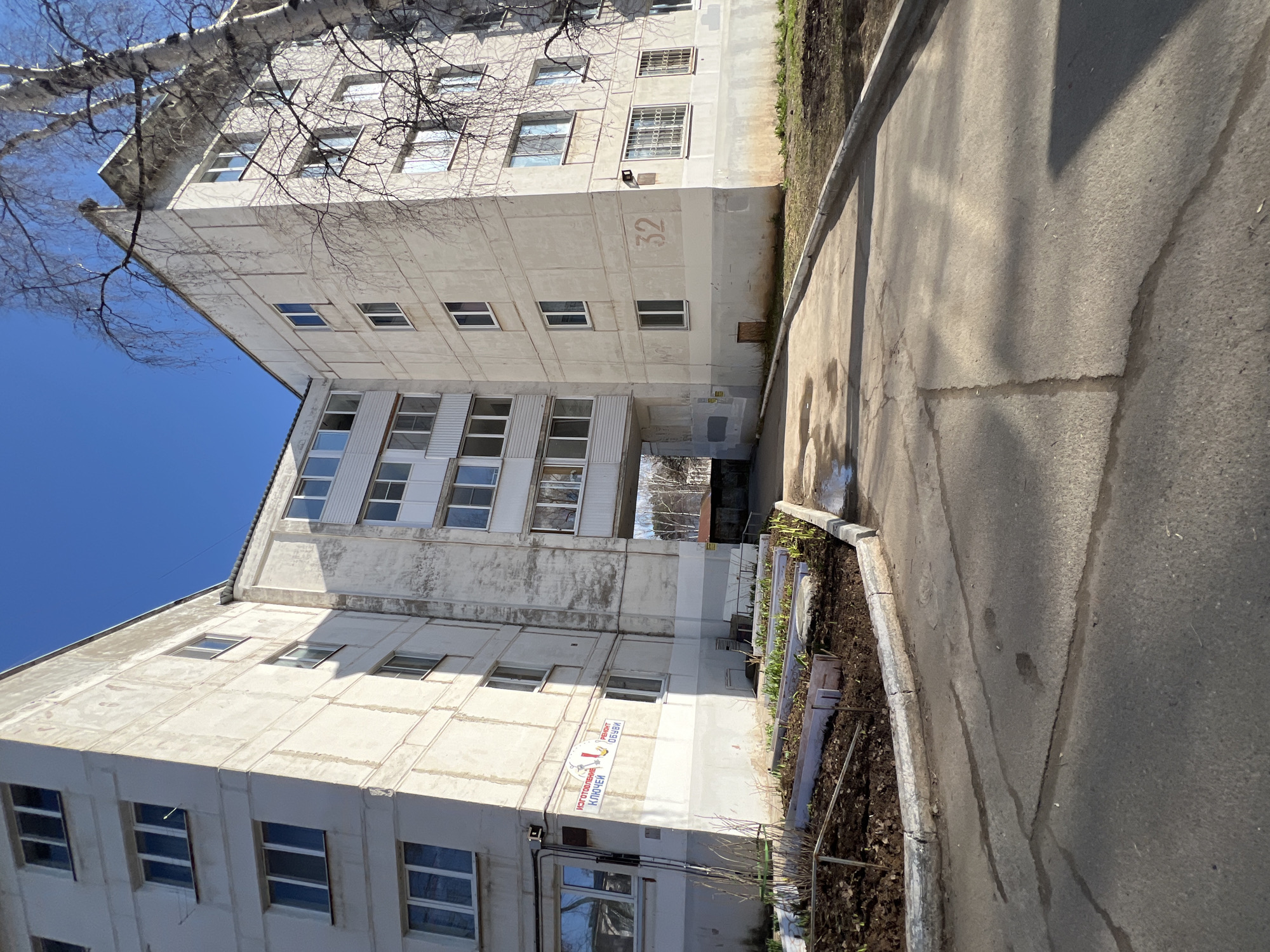 A medium shot depicts a Soviet-era concrete apartment building in Chaikovsky, Russia. The building has multiple stories with rows of windows. "32" is painted on the building's facade, indicating the building number. A pedestrian walkway runs alongside the building, showing cracks and wear. A small flowerbed or garden area is visible between the walkway and the building. The sky is clear and blue. Bare tree branches extend into the upper part of the frame. A sign on the building is in Russian, indicating a business making keys. The image is slightly rotated counter-clockwise.