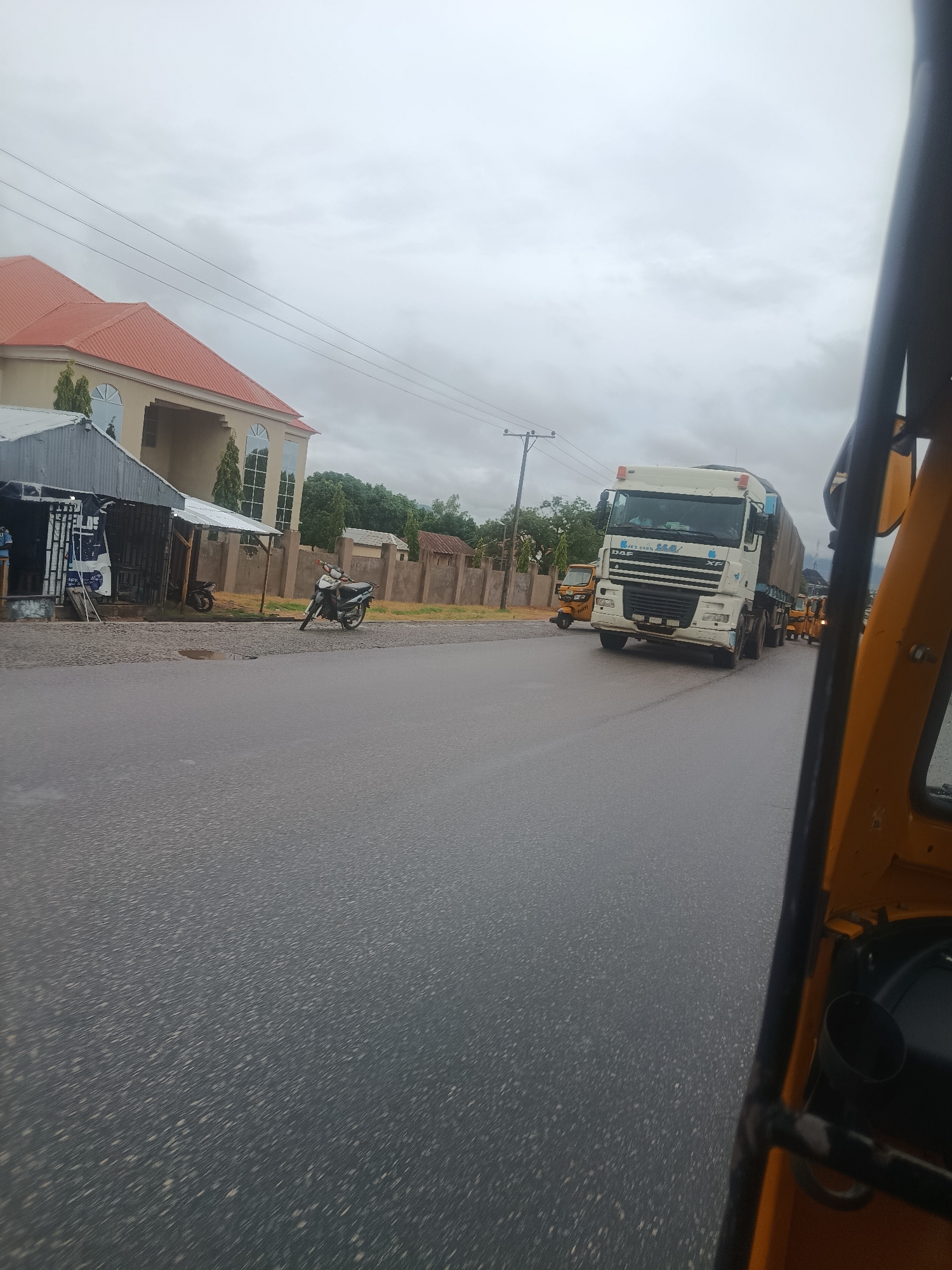 The image captures a view from a three-wheeled vehicle, likely a rickshaw, on a wet road in Jalingo, Nigeria. A large white truck with a canvas cover is driving on the road, followed by several yellow tricycles. In the background, there is a large building with a red roof and arched windows, a fence, and some trees. A motorcycle is parked on the side of the road near a small structure. The sky is overcast, suggesting cloudy or rainy weather. The road surface appears to be asphalt with visible patches and some puddles, indicating recent rain. The perspective suggests the photo was taken while the vehicle was in motion.