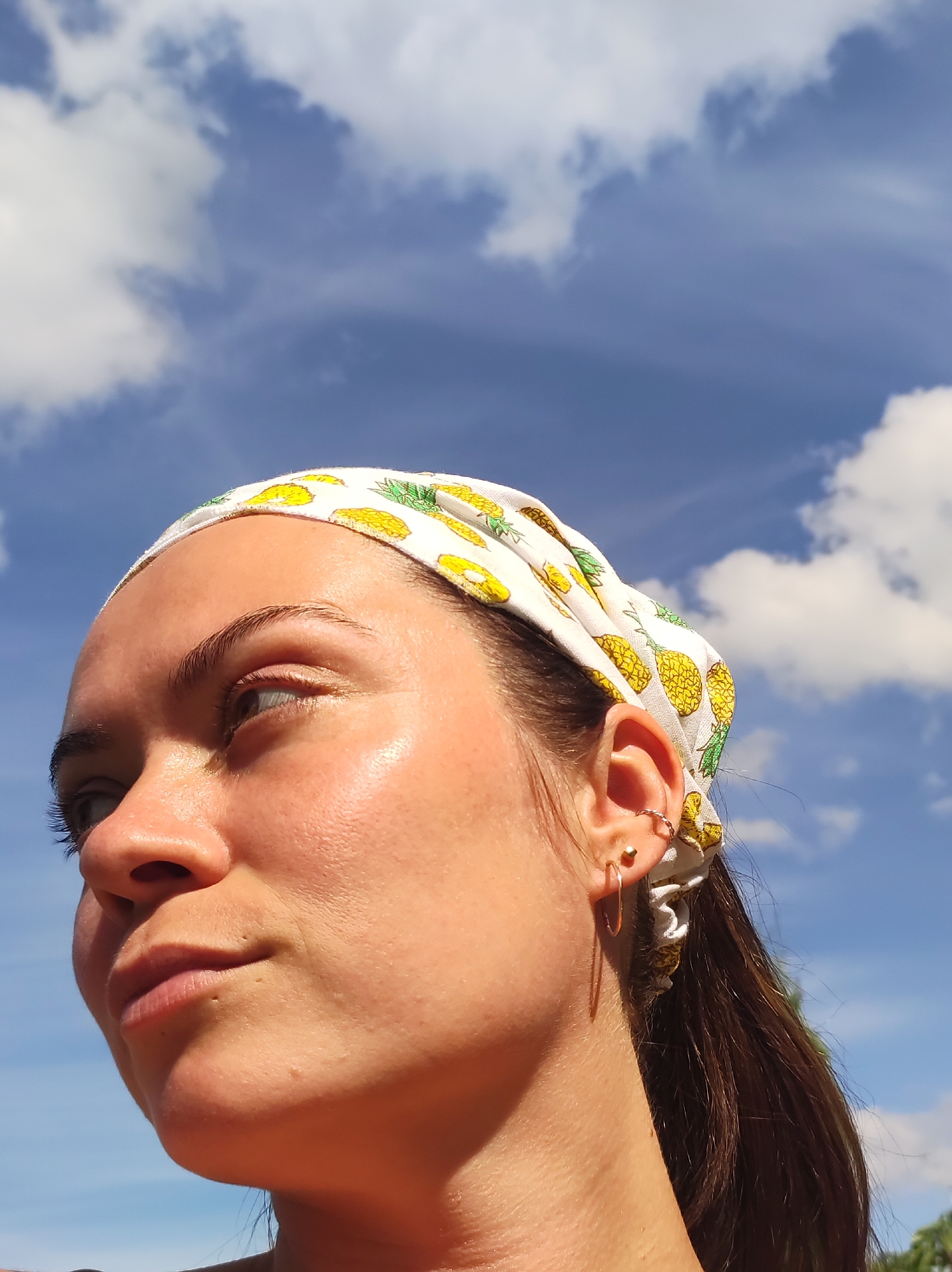 A close-up, profile shot shows the right side of a young woman's face, looking upwards and to her left. She is wearing a white bandana with a pineapple print, which is tied around her head. Her dark hair is pulled back. She has an earring in her upper ear, a small gold stud, and a thin gold hoop earring in her lower earlobe. Her skin appears smooth and slightly dewy. The background is a bright blue sky with scattered white clouds. The lighting suggests it is daytime, possibly outdoors. There are no city landmarks or urban elements visible.