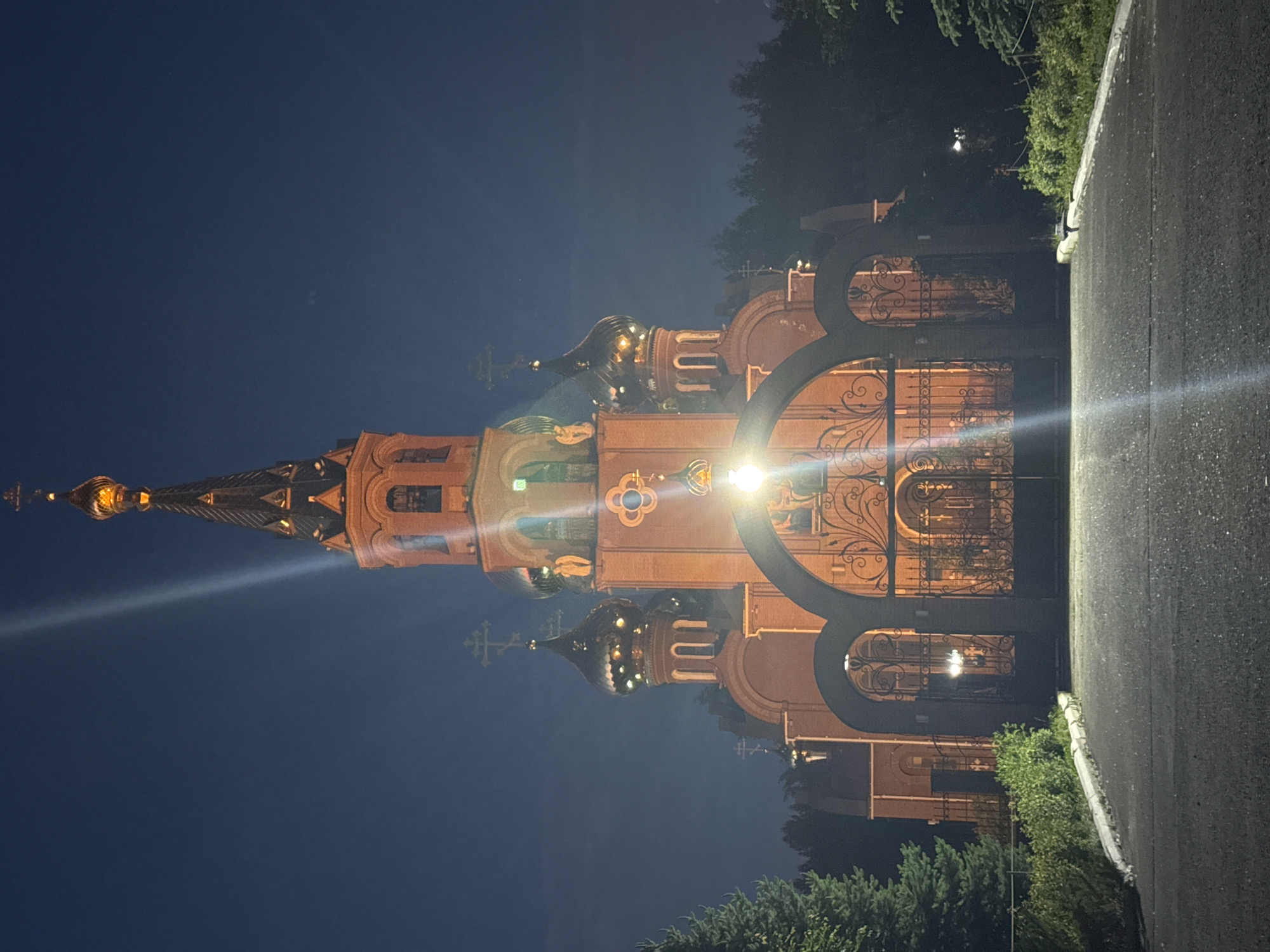 The image captures an illuminated Eastern Orthodox church at night, set against a dark blue-black sky. The prominent subject is a grand church building, constructed from reddish-brown brick or stone. It features a tall central spire culminating in a golden onion dome topped with a cross, surrounded by several other smaller golden onion domes, each also bearing a cross. The church exhibits ornate architectural details, including arched windows and decorative cornices.
In front of the church, a large, elaborate black metal gate with intricate scrollwork and arched designs is visible, suggesting an entry point to the religious complex. A bright artificial light source, likely a streetlight, positioned to the right of the gate, casts a prominent lens flare across the scene, illuminating parts of the gate and the church facade.
Along the right side of the frame, a paved surface, possibly a road or path, runs parallel to the church property. Dark green trees and bushes are visible around the church and along the foreground, adding natural elements to the nocturnal setting.
The time of day is clearly night, characterized by the dark sky and the warm glow of artificial lighting on the church, which highlights its architectural beauty. The weather appears clear. The distinct architectural style, particularly the onion domes and crosses, points to an Orthodox Christian church, consistent with the given location context of Chaikovsky, Russia. No people or visible text are present in the image, which depicts a static, serene scene.