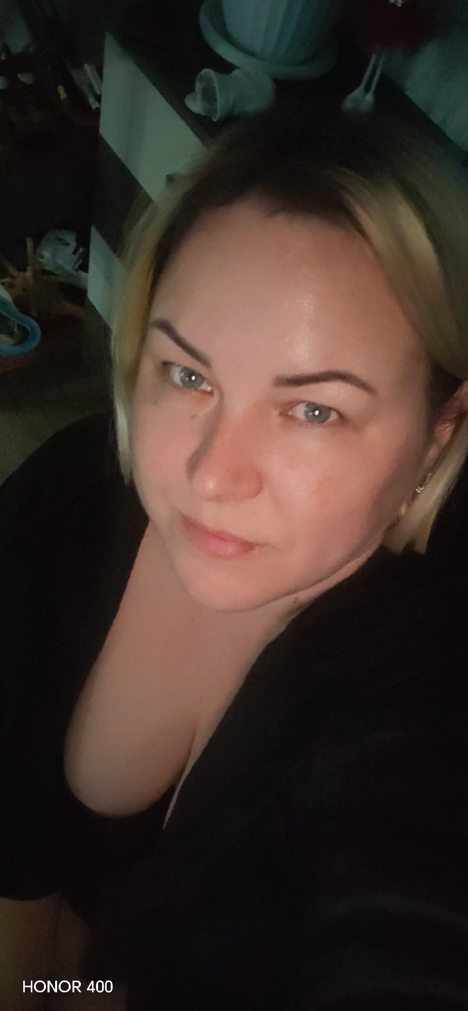 A woman with blonde hair and blue eyes poses for a selfie in what appears to be an indoor setting. She is wearing a black top. The background is dimly lit and cluttered with various objects, including a white planter. The location is unknown, and there is no evidence to confirm it is in the city of Noviy, Russia. The image includes the text "HONOR 400" at the bottom.