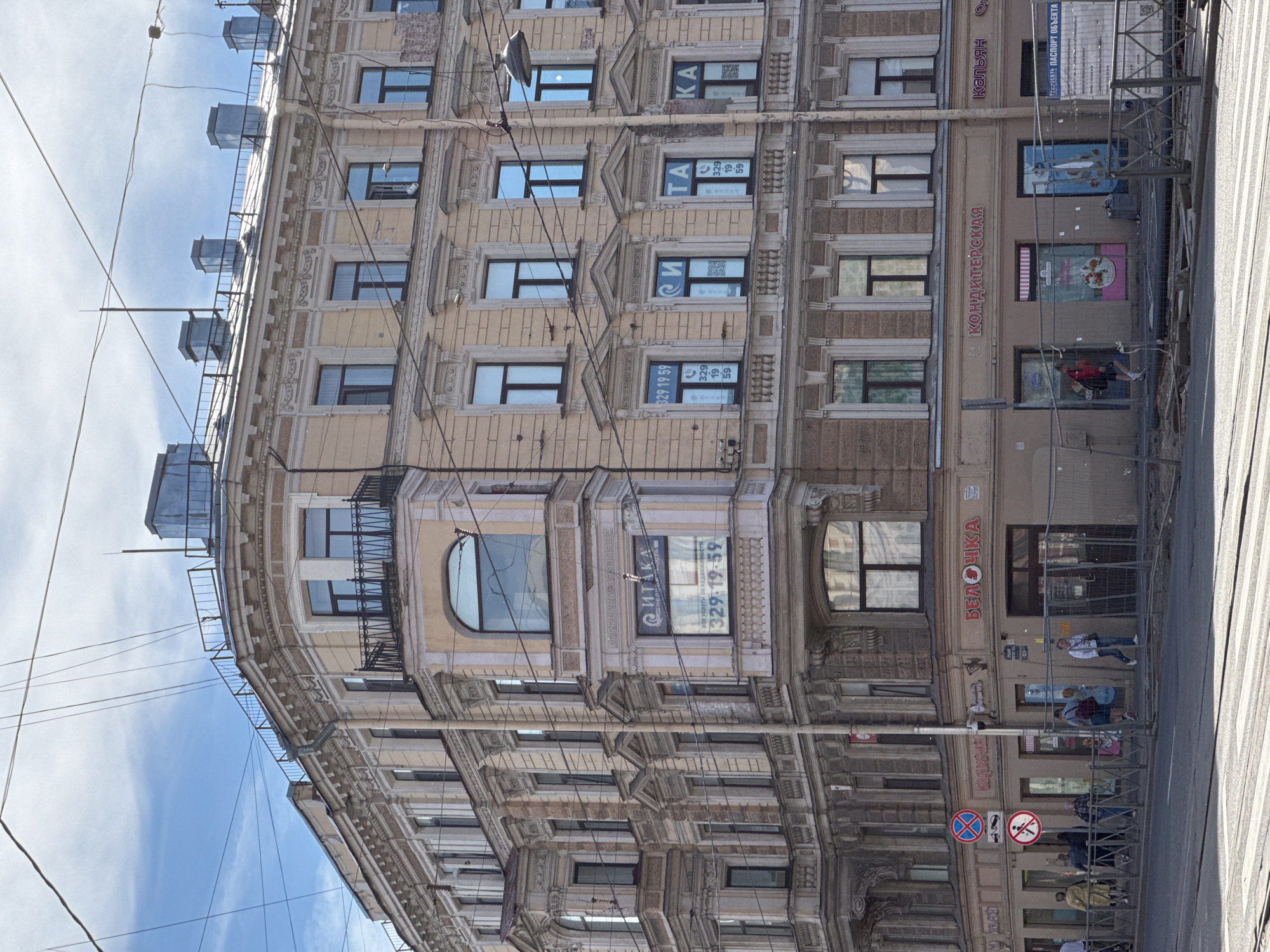 A multi-story building with ornate classical architecture, painted in shades of yellow and beige, stands on a city street in Saint Petersburg, Russia. The building features large windows, some with balconies, and intricate decorative elements along its facade. Overhead, a network of electrical wires crisscrosses the sky, which is partly cloudy.
The ground floor of the building houses several storefronts, some of which are under renovation, indicated by temporary fencing and construction materials. Signs above the shops display various text in Russian, including "БЕЛОЧКА" (Belochka) and "КОНДИТЕРСКАЯ" (Confectionery). People are visible walking along the sidewalk in front of the shops, and some are seen crossing the street at a pedestrian crossing.
The street in front of the building has tram tracks. The overall scene depicts an active urban environment with a mix of historical architecture and modern street life.
