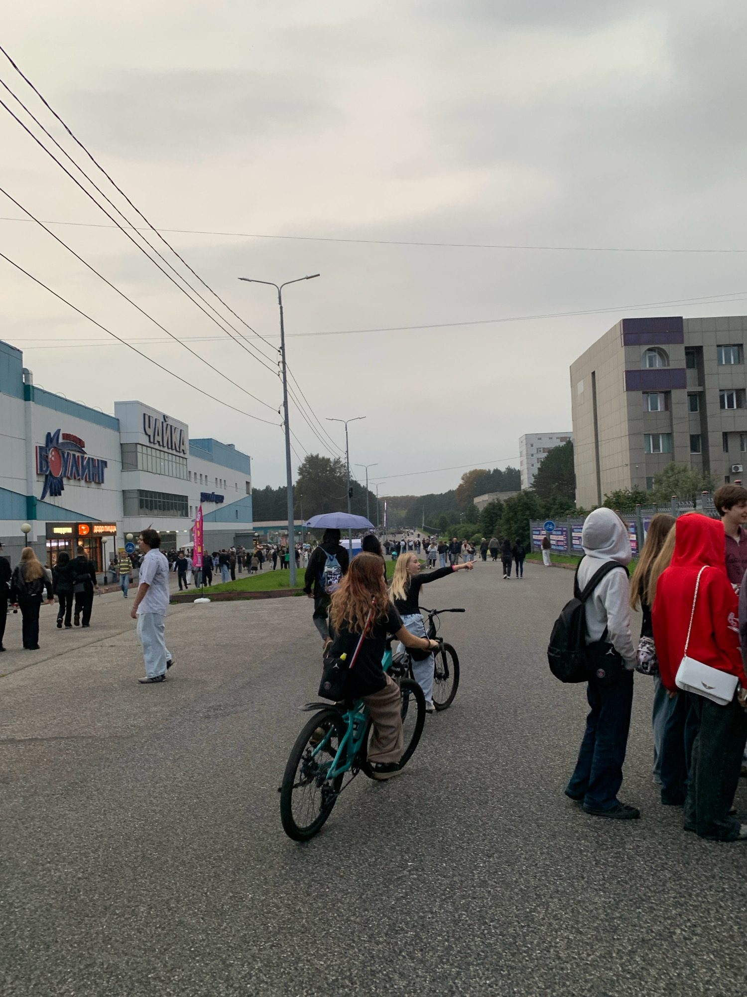 The image depicts an outdoor scene with a group of people gathered on an asphalt surface, likely a street or parking lot, in Prokopyevsk, Russia. A large, modern building with "ЧАЙКА" and "БОУЛИНГ" signage in Russian is visible on the left, suggesting a commercial area. In the background, more buildings, trees, and utility poles with power lines are present under an overcast sky.
Several individuals are engaged in various activities. In the foreground, two young women are on bicycles. One, wearing a black top and brown pants, is on a turquoise bicycle, looking forward. The other, with blonde hair and a black top, is also on a bicycle and appears to be pointing. To their right, a group of people are standing and talking, including a young person in a grey hooded sweatshirt with a black backpack, and another in a bright red hoodie holding a white purse. Further to the right, two young men are visible, one in a maroon shirt and another in a dark shirt.
In the middle ground, a person in a white medical uniform walks away from the camera. Other individuals are spread out, some walking and some standing, in front of the commercial building. An umbrella is visible over the head of a person in a black jacket. The overall atmosphere suggests a casual gathering or event.