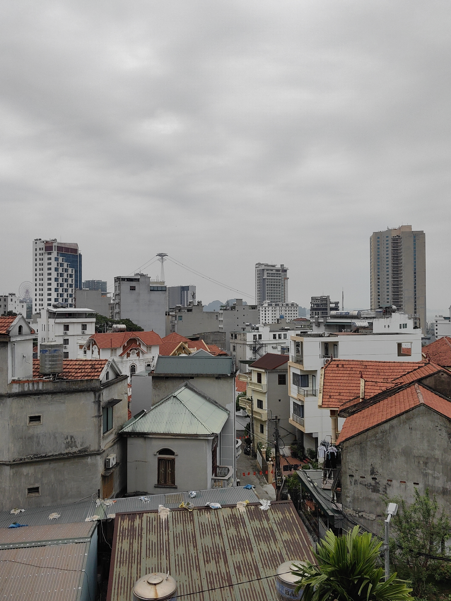 The image captures a view of Bai Chay Ward, Vietnam, showcasing a dense urban environment under a cloudy sky. The foreground is dominated by rooftops of varying materials, including concrete, metal, and red tiles. A network of streets and buildings is visible, revealing a mix of residential and commercial structures. In the background, several high-rise buildings punctuate the skyline, including a building with a Ferris wheel. A tall cable car tower can also be seen. A street with red flags lining it is also visible. Laundry hangs from a building on a line, and a palm tree grows.