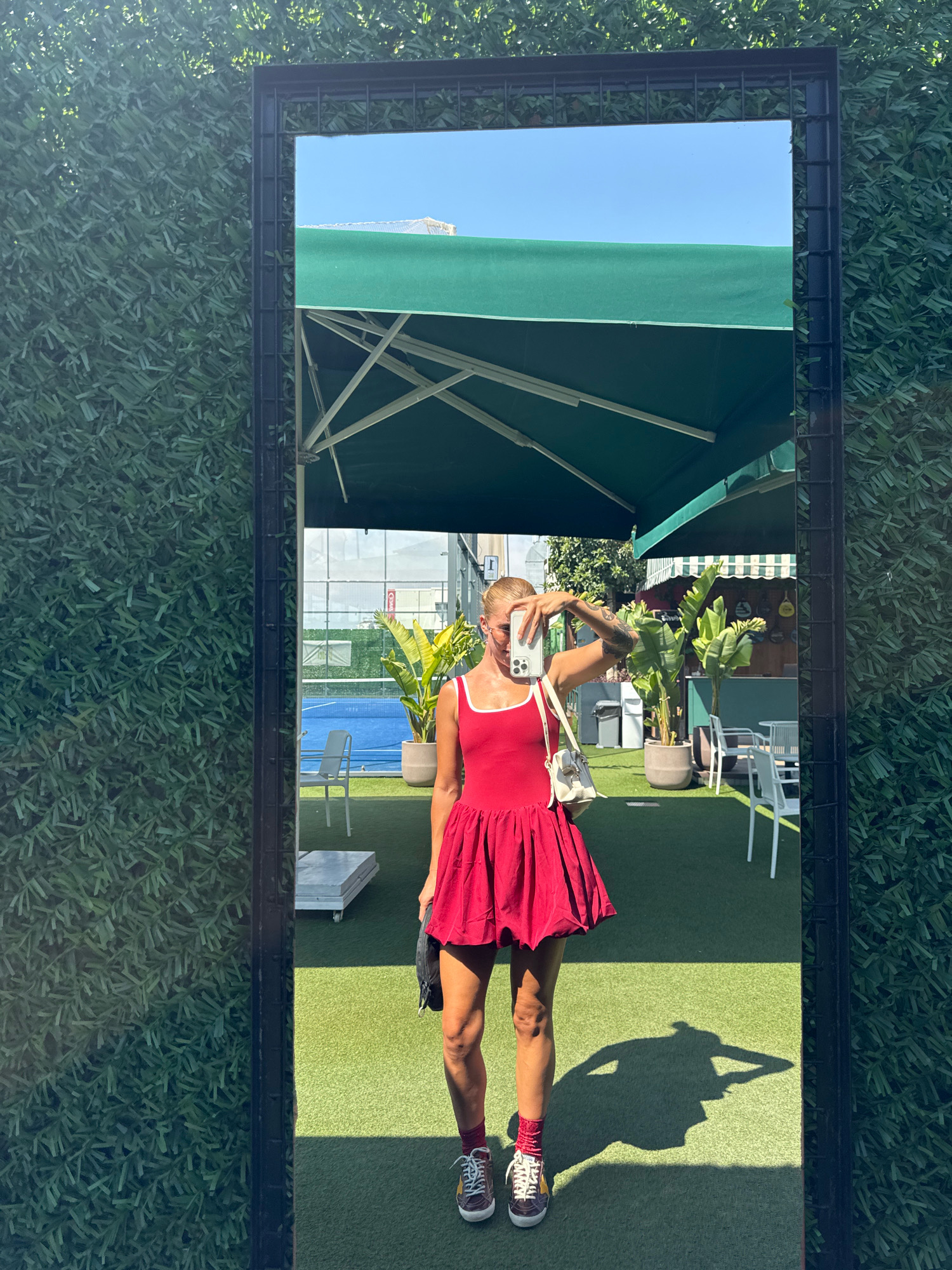 A woman is taking a selfie in a mirror, wearing a red dress, white socks, and sneakers. She is standing on a green turf surface, with a shadow of herself cast on the ground. The reflection in the mirror shows a tennis court, palm trees, and outdoor seating. The background is a vertical green hedge. The image is taken in Antalya, Turkey.