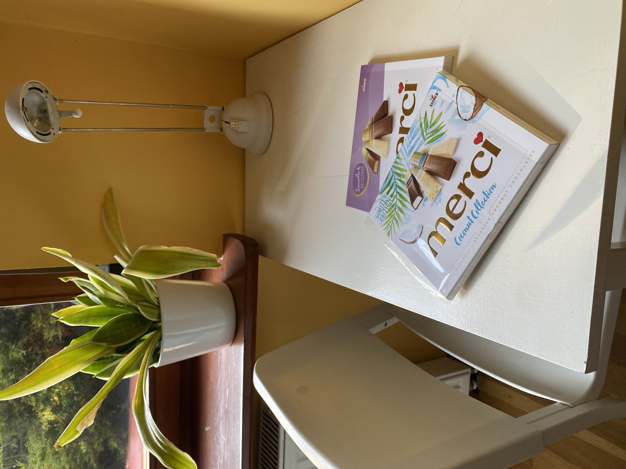 An indoor scene shows a white table with two boxes of Merci assorted coconut chocolates on it. The box in the foreground is white with gold lettering and illustrations of palm leaves and coconut. The box behind it is purple with an illustration of different chocolate bars. To the left of the table is a white chair. Behind the table is a yellow wall. On the left side of the frame, a plant in a white pot sits on a dark wooden windowsill. Outside the window, green foliage is visible. To the right of the plant is a white desk lamp. The scene appears to be in an apartment in Wrocław, Poland.