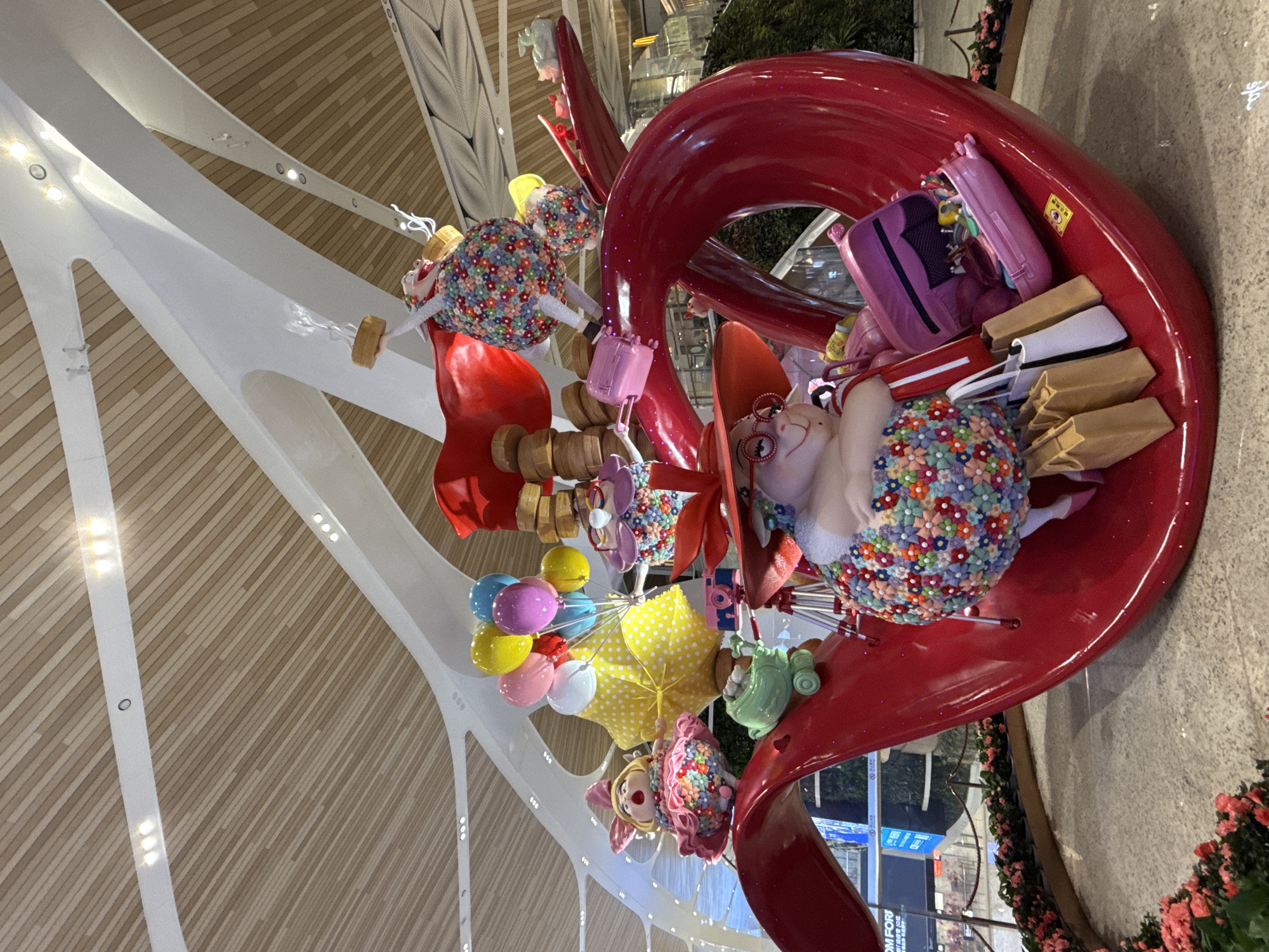 The image shows a whimsical art installation within an interior space, potentially an airport terminal, given the presence of structural beams and distant signage. The installation features oversized figures and objects arranged in a large, red, swirling structure. The figures are cartoonish, with colorful floral patterns and exaggerated features. One figure wears an orange hat and glasses. Accessories like shopping bags, a purple suitcase, a polka-dotted umbrella, and balloons are part of the display. The terminal's interior features wooden panels. Based on the location information, this is likely located in the Baiyun District, China.