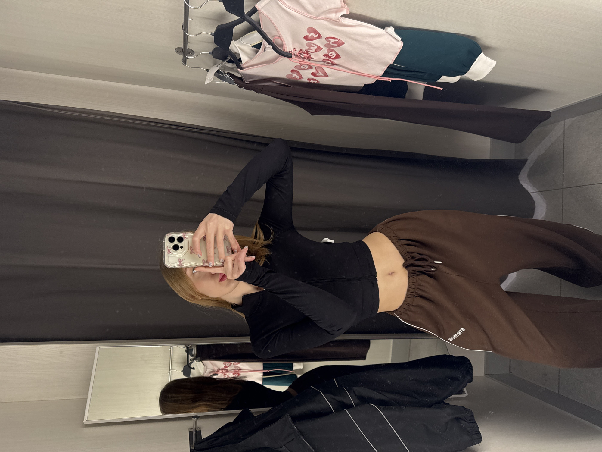 A young female, with long blonde hair, red lipstick, and manicured nails, is taking a mirror selfie with a smartphone inside a changing room in Rostov-on-Don, Russia. She is wearing a black long-sleeved zip-up crop top and brown drawstring sweatpants with a white side stripe displaying text, revealing her navel. The smartphone, held in her right hand, features a clear case with a decorative pattern. To her left, a full-length mirror reflects her image and a dark jacket with white stripes hanging on a wall hook. To her right, a metal clothing rack holds various garments, including a light pink short-sleeved top with red heart graphics and pink lacing, alongside other items like dark green and brown clothing. The changing room walls are light-colored, and the floor is tiled. A dark grey fabric curtain serves as a partition.