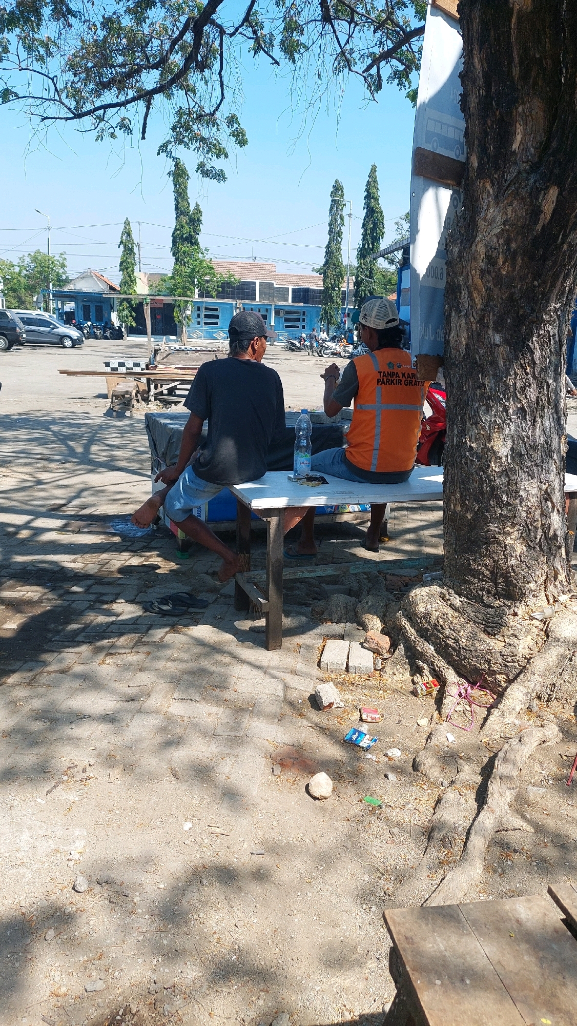 Two men are seated at a table outdoors on a sunny day in Sidoarjo, Indonesia. One man, wearing a dark t-shirt and a black baseball cap, sits with his legs crossed, facing away from the camera. The other man, wearing an orange reflective vest over a t-shirt and a baseball cap, is seated on the same bench, facing the first man. He appears to be holding something in his hands. A bottle of water and a small packet are on the table between them. They are seated near the base of a large tree with exposed roots, on a surface of dirt and scattered stones and debris. In the background, there are buildings with blue accents, parked cars, and motorcycles, suggesting a public area or parking lot. Sunlight casts shadows through the leaves of trees overhead onto the ground.