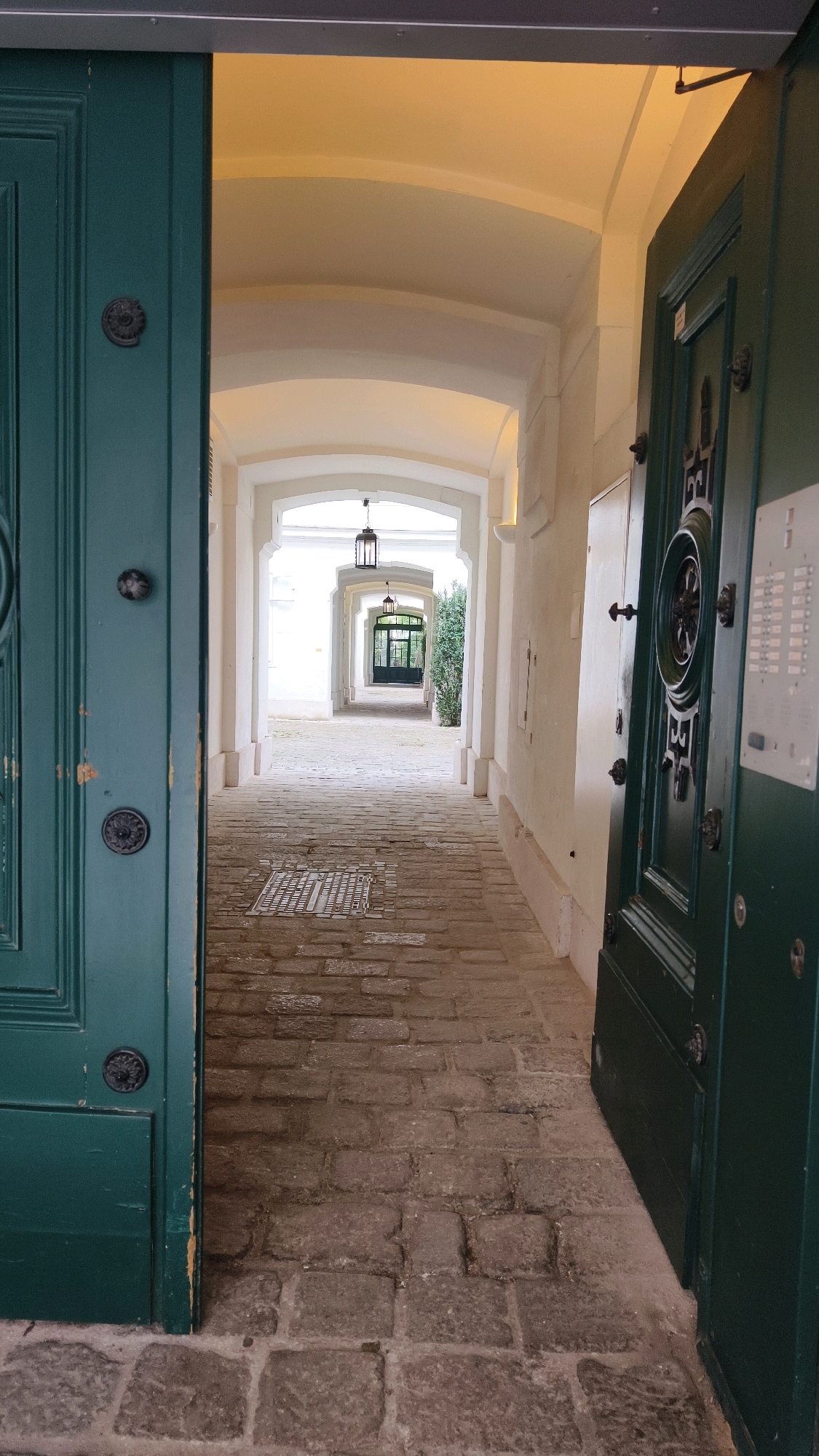 An open green wooden door reveals a cobblestone pathway leading through an arched courtyard in Vienna, Austria. The pathway is lined with white stucco walls and is illuminated by overhead lights in antique-style lanterns. The door on the right is also green and appears to be a main entrance, featuring decorative metalwork and a modern intercom system. The scene is viewed from an exterior perspective, looking into the courtyard.